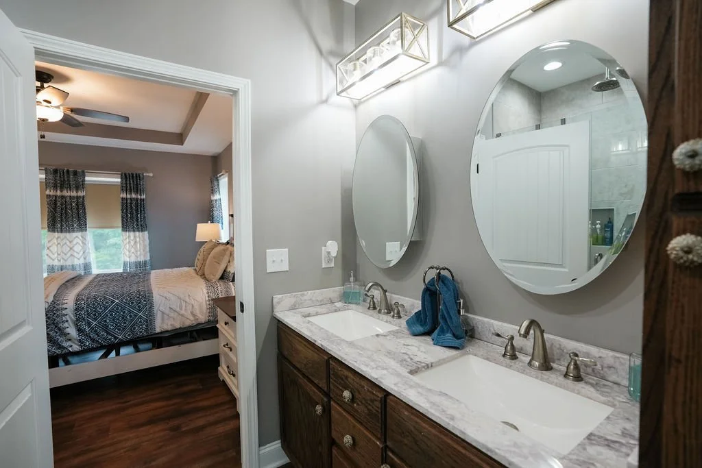 Modern bathroom with dual sinks and round mirrors, gray walls, and view into a bedroom with a bed and ceiling fan.
