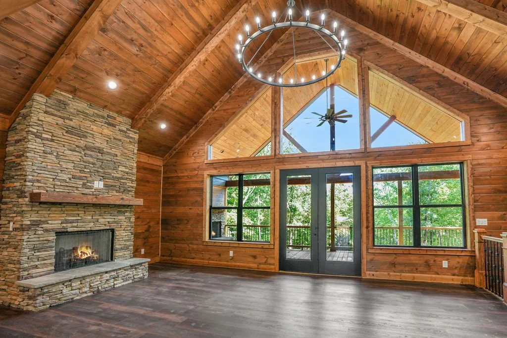 Modern rustic living room with vaulted wooden ceiling, large stone fireplace, circular chandelier, glass doors, and large windows overlooking greenery.