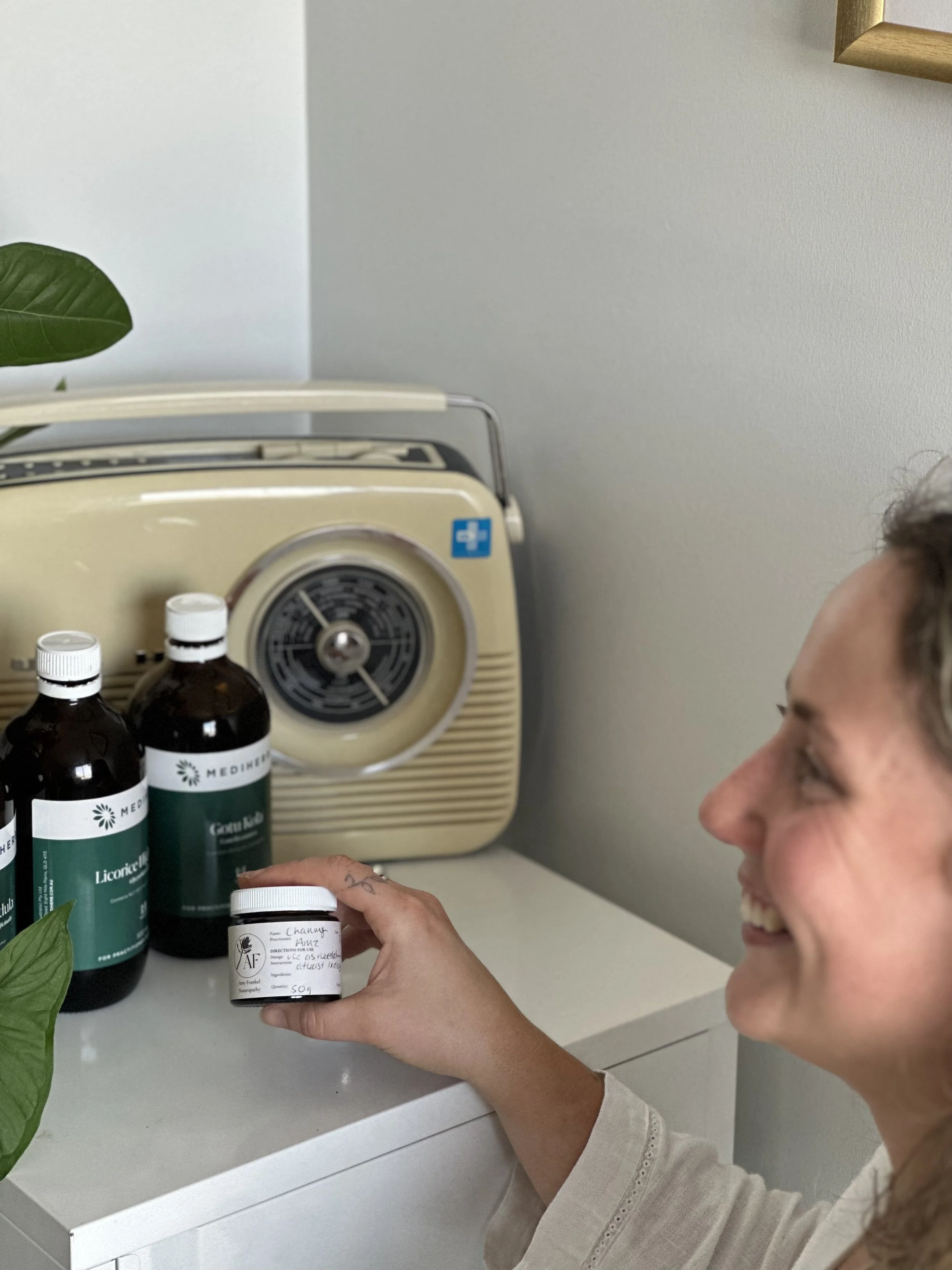 Smiling woman holding a small jar labeled 'Chama' with some handwritten notes, standing next to bottles of medicine and a vintage photo radio on a white cabinet.