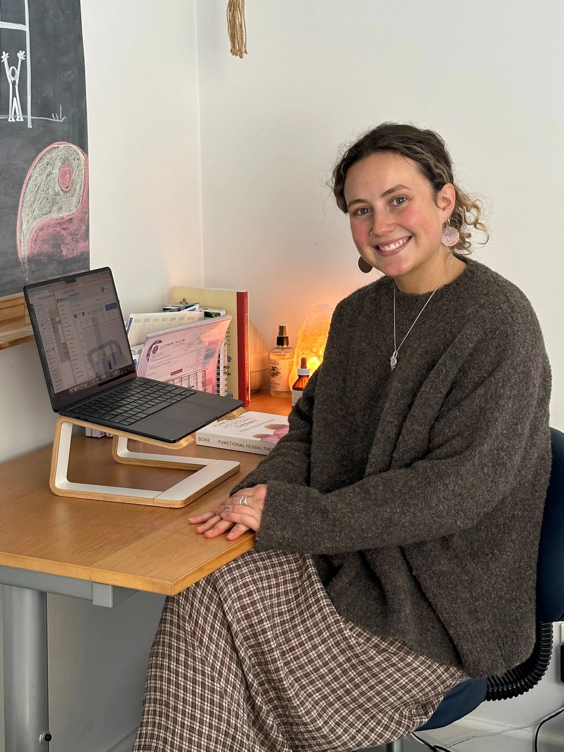 A woman sitting at a desk with a laptop, notebooks, and various herbal products, smiling at the camera in an office space.