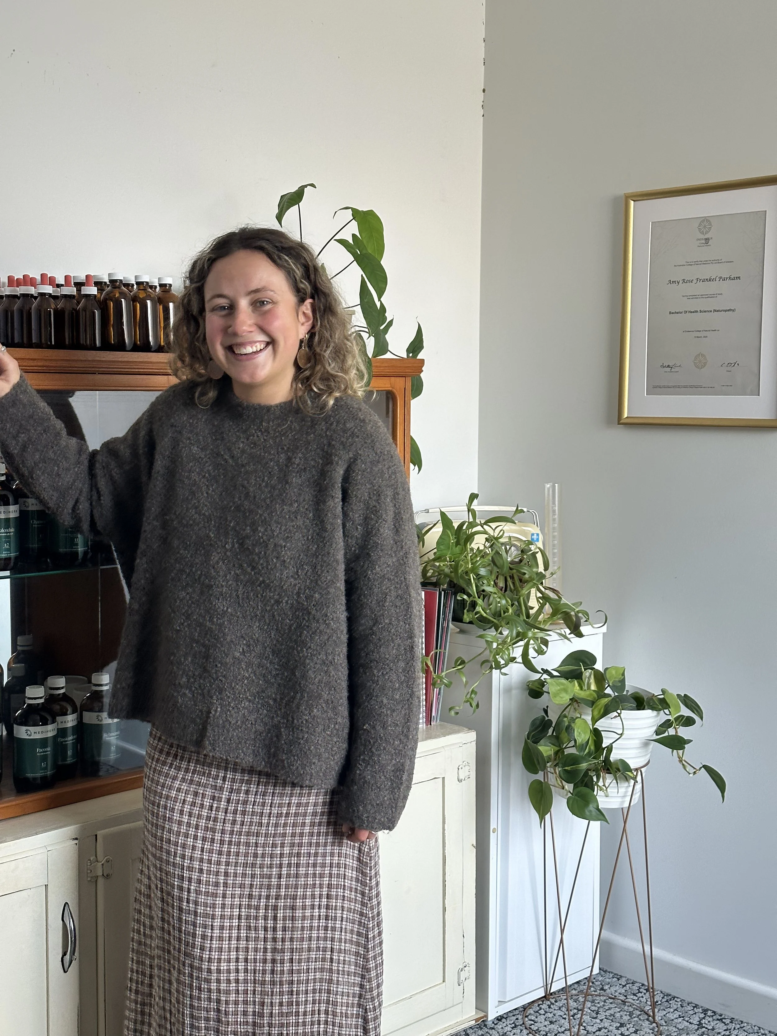A woman with curly hair smiling, wearing a brown sweater and a plaid skirt, standing indoors next to potted plants and a framed diploma.