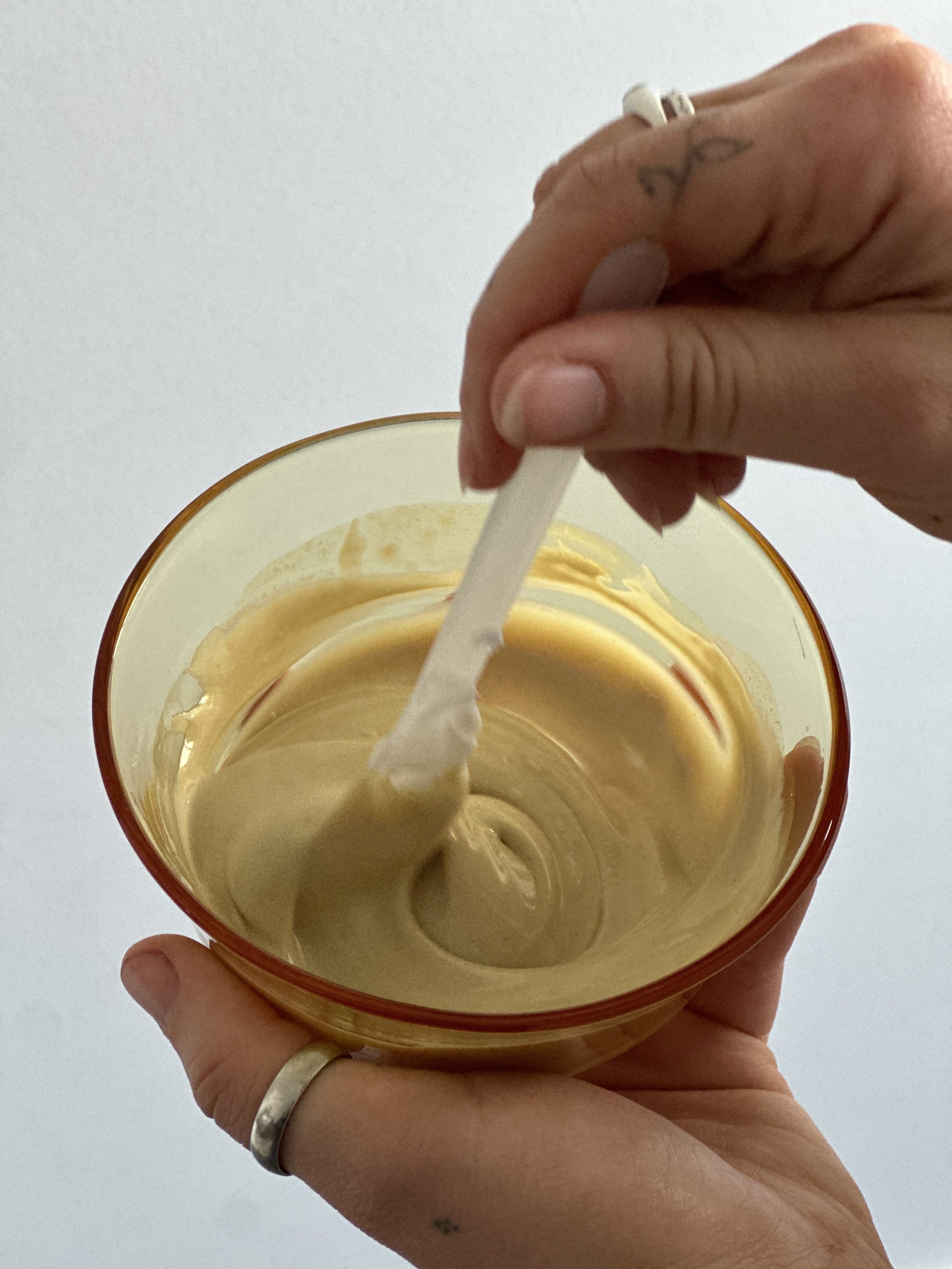 Person holding a glass bowl of yellowish caramel or fudge, stirring it with a white plastic spatula.