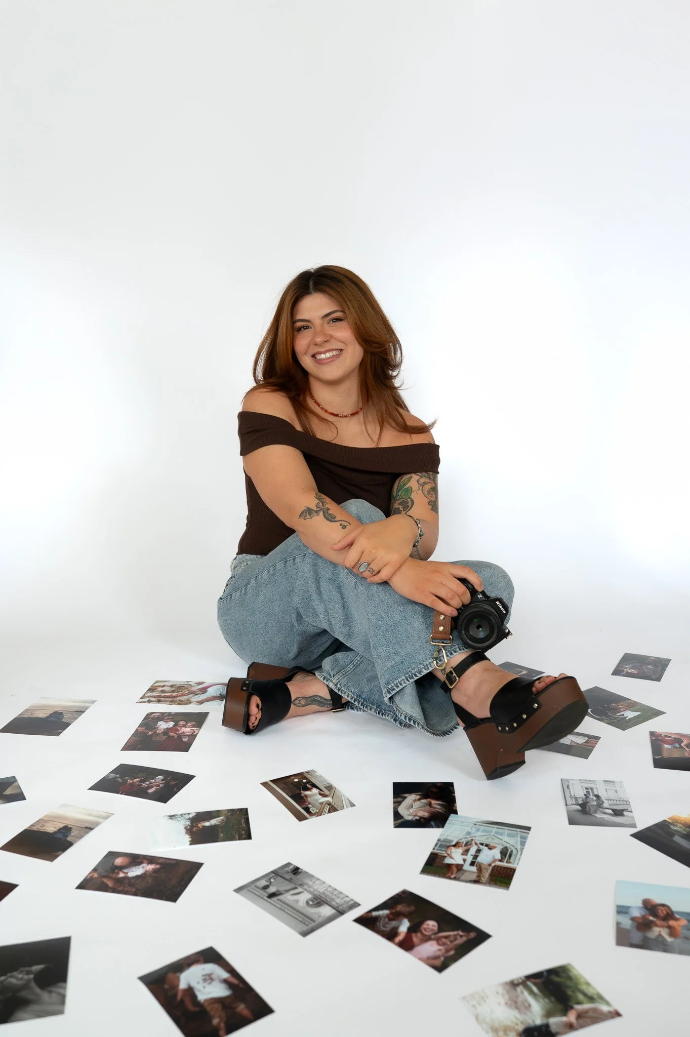 A young woman with brown hair sitting on a white floor, surrounded by scattered photographs. She is smiling, wearing a black off-the-shoulder top, light blue jeans, and chunky black sandals. There is a camera in her lap and some photographs in front of her.