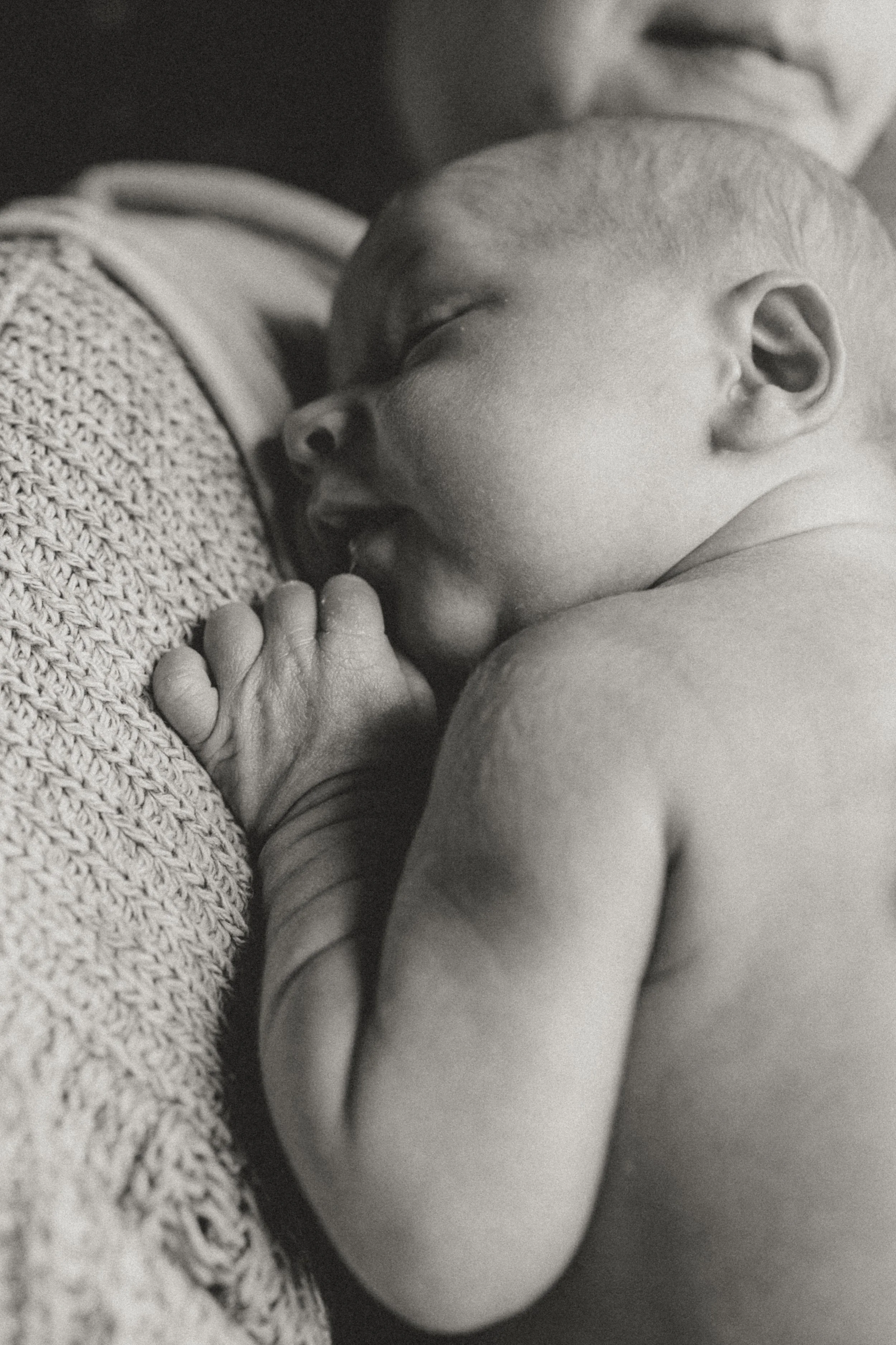 Close-up black and white photo of a person holding a sleeping baby on their shoulder. The baby has a bald head, closed eyes, and is resting peacefully with its hand near its mouth. The person is wearing a textured sweater and appears to be giving gentle support.