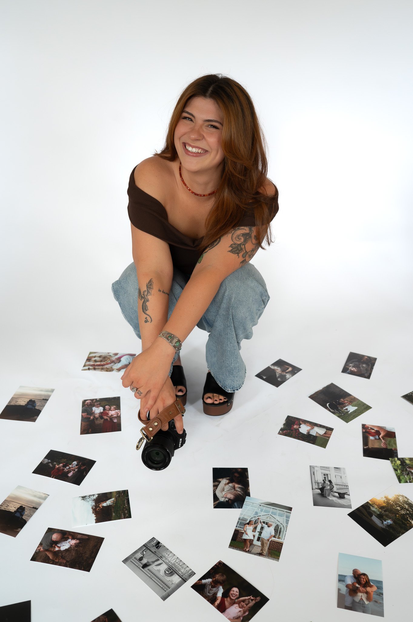 Young woman with tattoos and shoulder-length hair smiling at camera, surrounded by scattered photographs on the floor, holding a camera, in a photography studio.