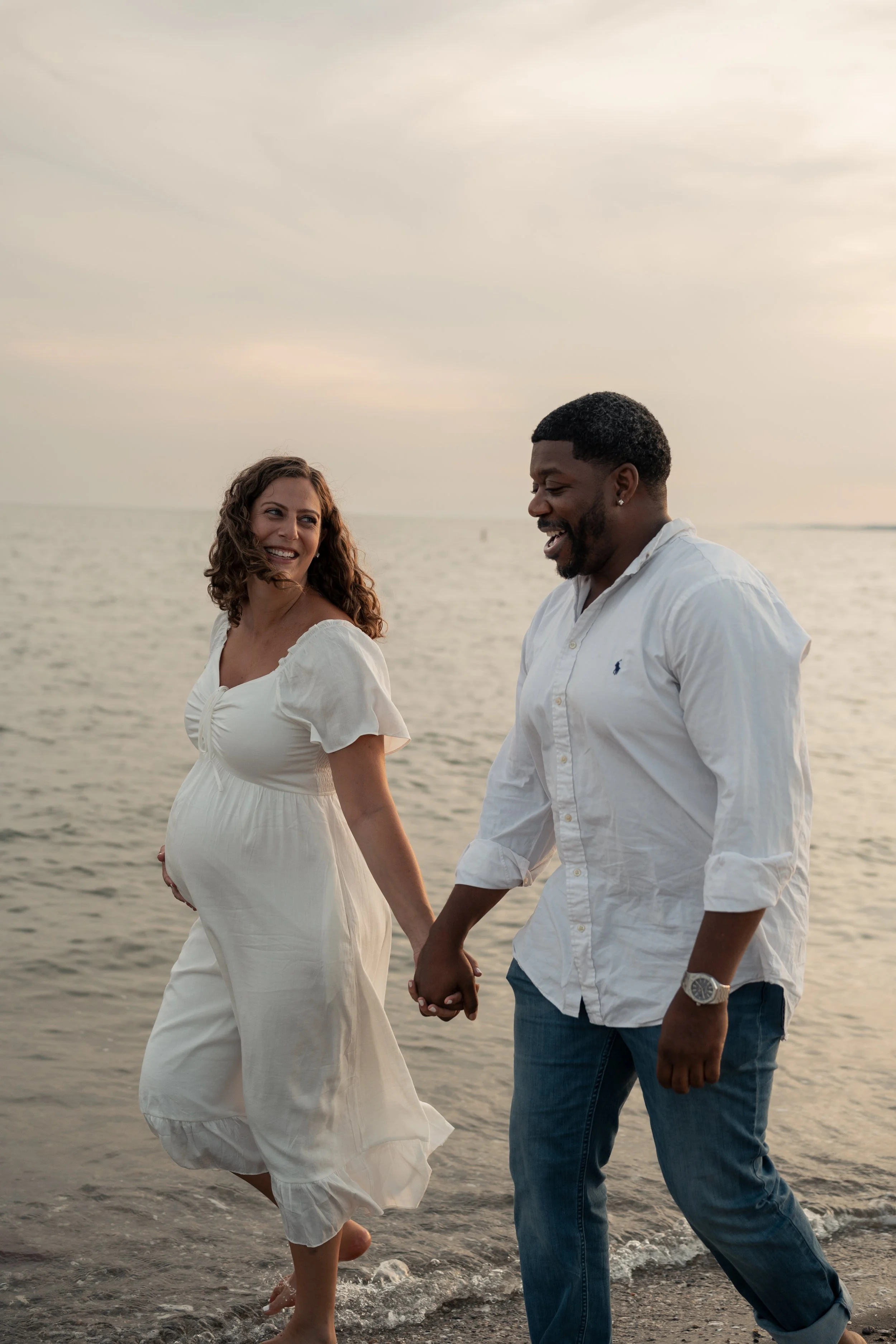 A pregnant woman and a man are holding hands and walking along the beach at sunset, smiling at each other.