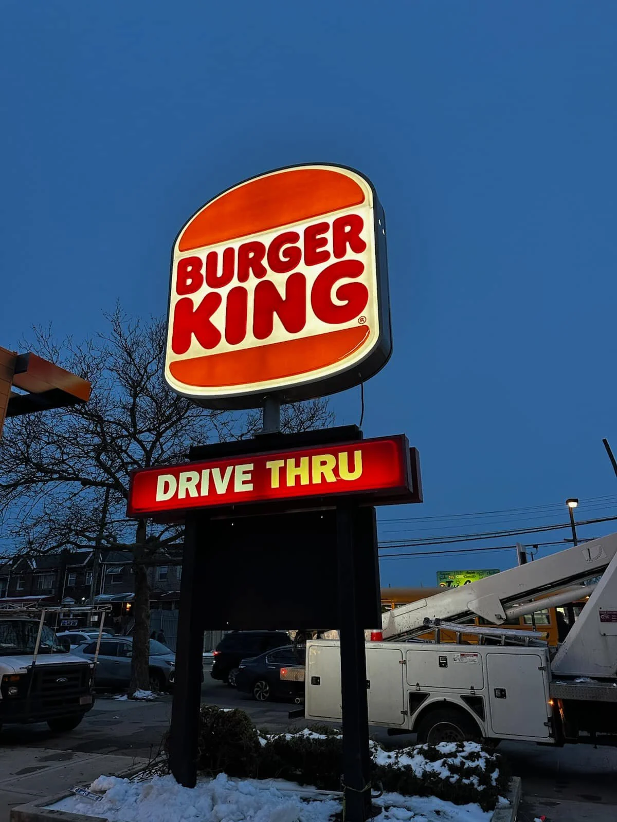 Burger King drive-thru sign at night with surrounding vehicles and snow on the ground.
