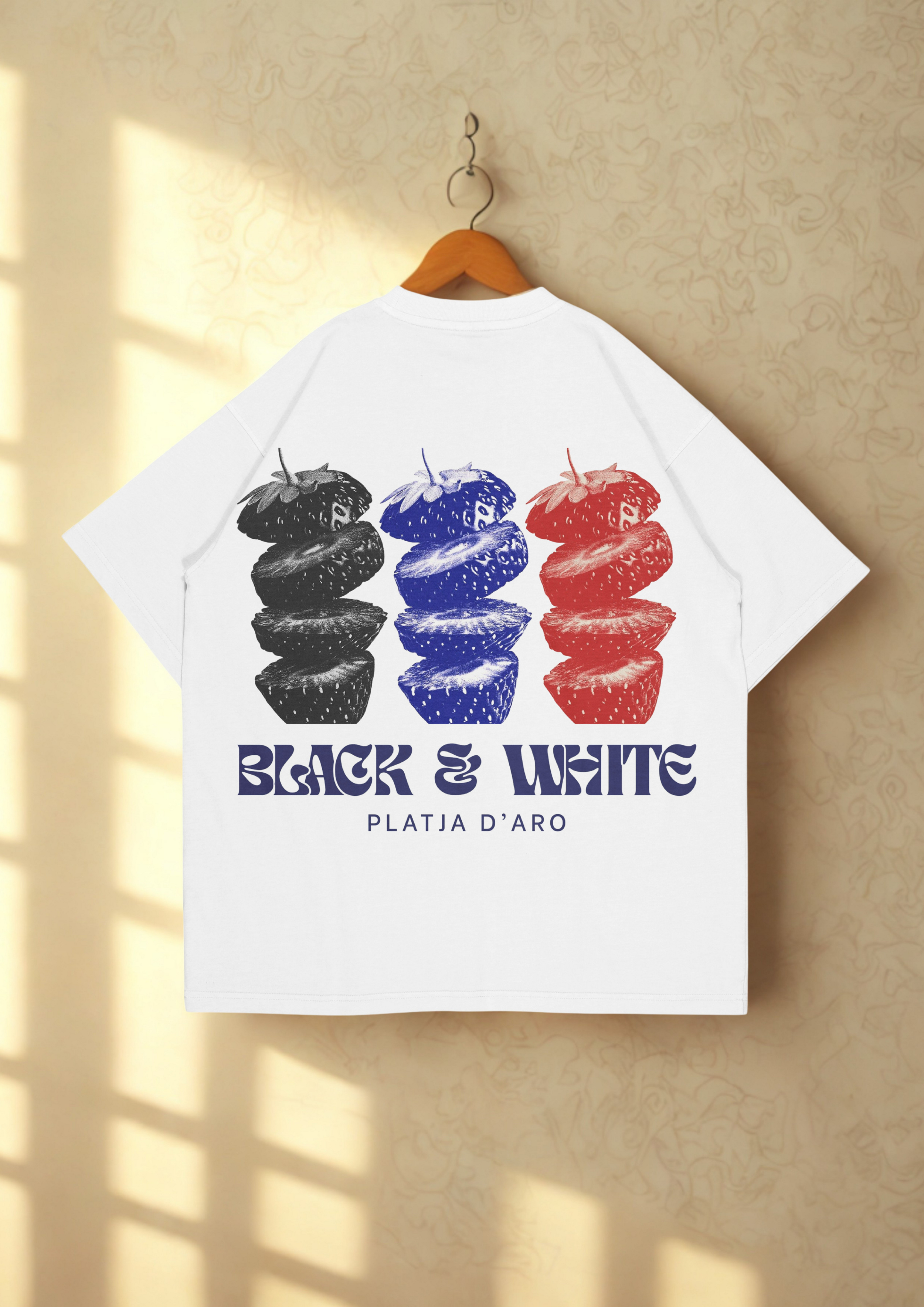 Camiseta blanca con diseño de tres fresas en colores negro, azul y rojo y texto que dice "Black & White" y "Plata D'Oro" debajo, colgada en un perchero de madera en una pared beige con patrón sutil.