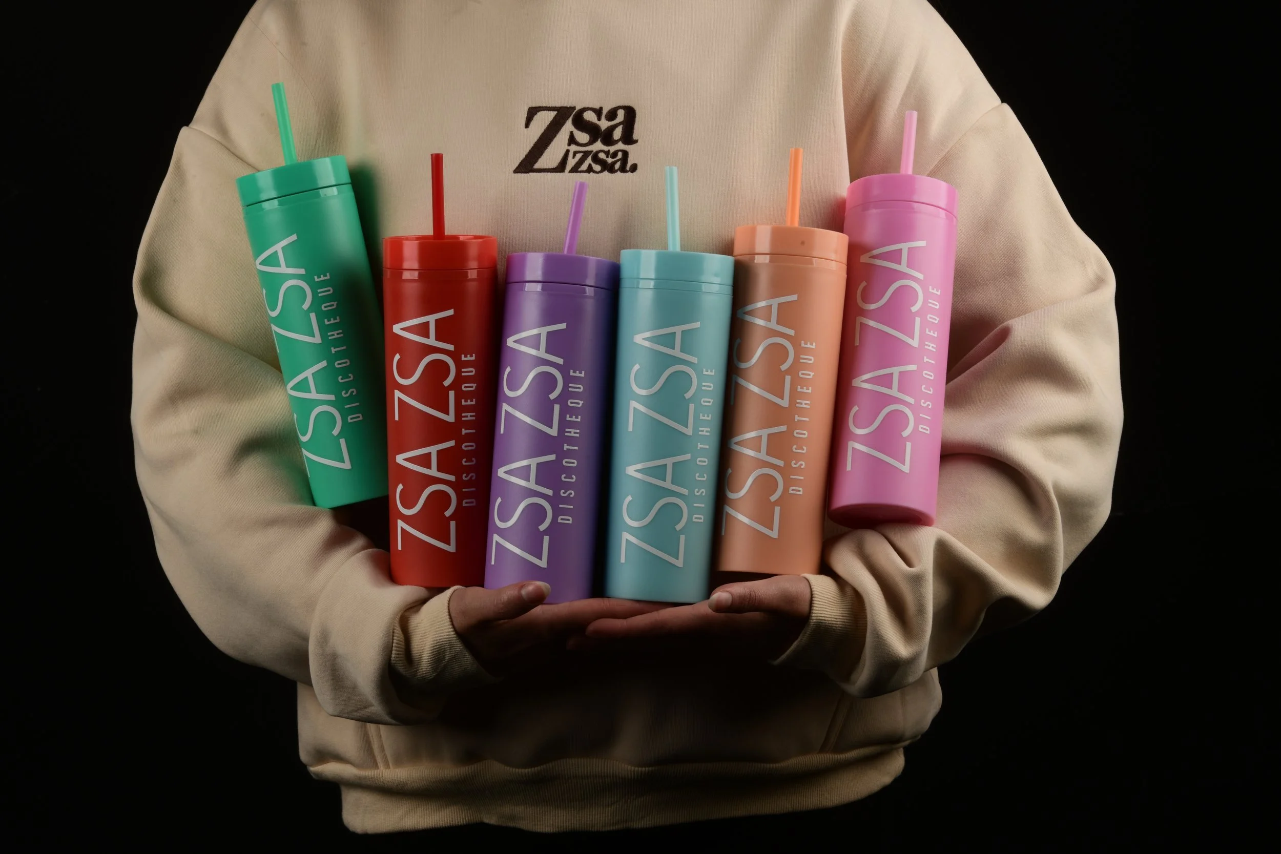 Una persona con sudadera beige sostiene varias botellas de plástico en colores pastel, cada una con una pajilla y el logo de Zsa Zsa Discuiseque en ellas.