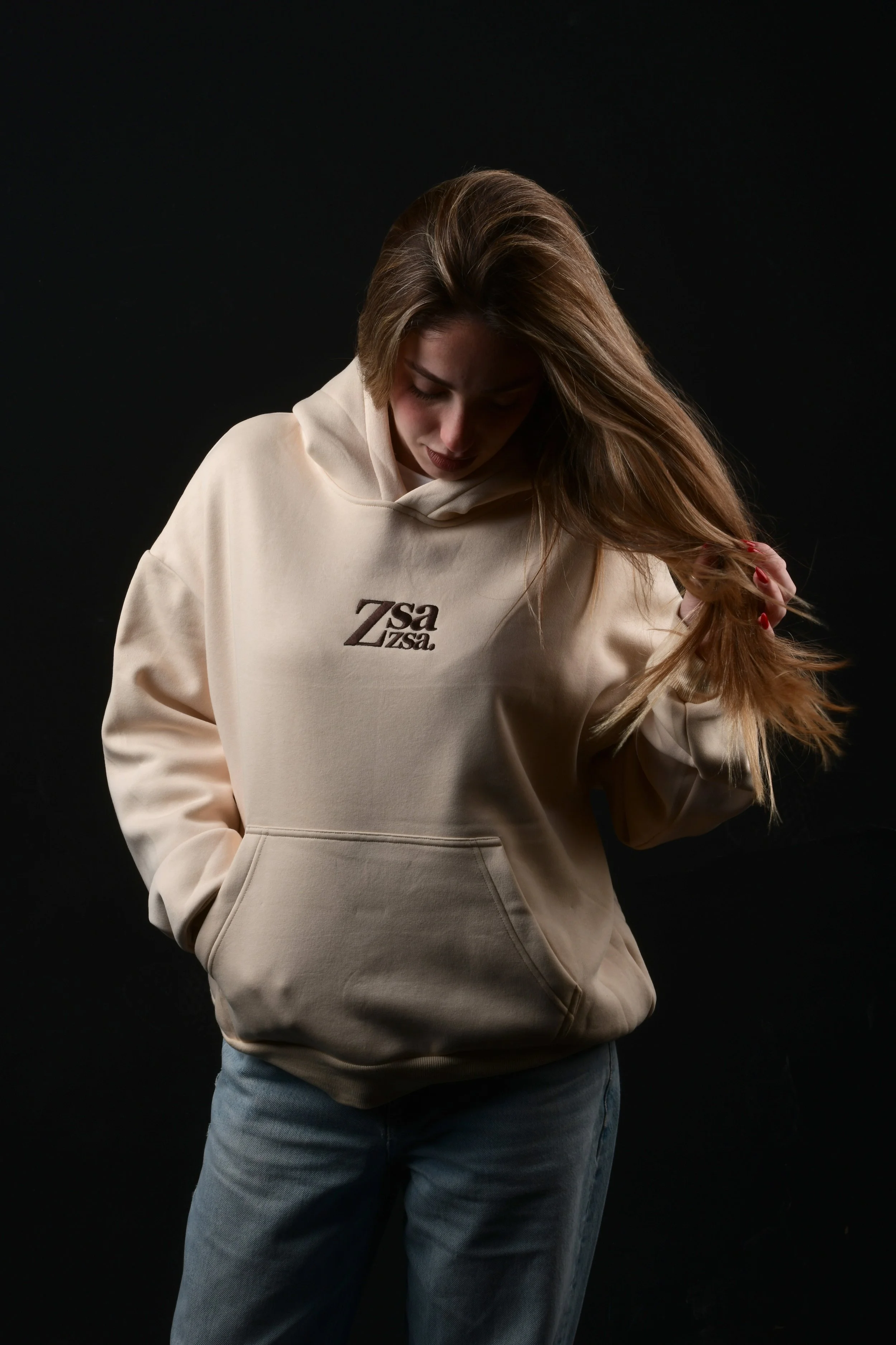 Mujer con sudadera beige que tiene texto que dice '7 s a Ziza' en un fondo negro, mirando hacia abajo y sosteniendo su cabello.