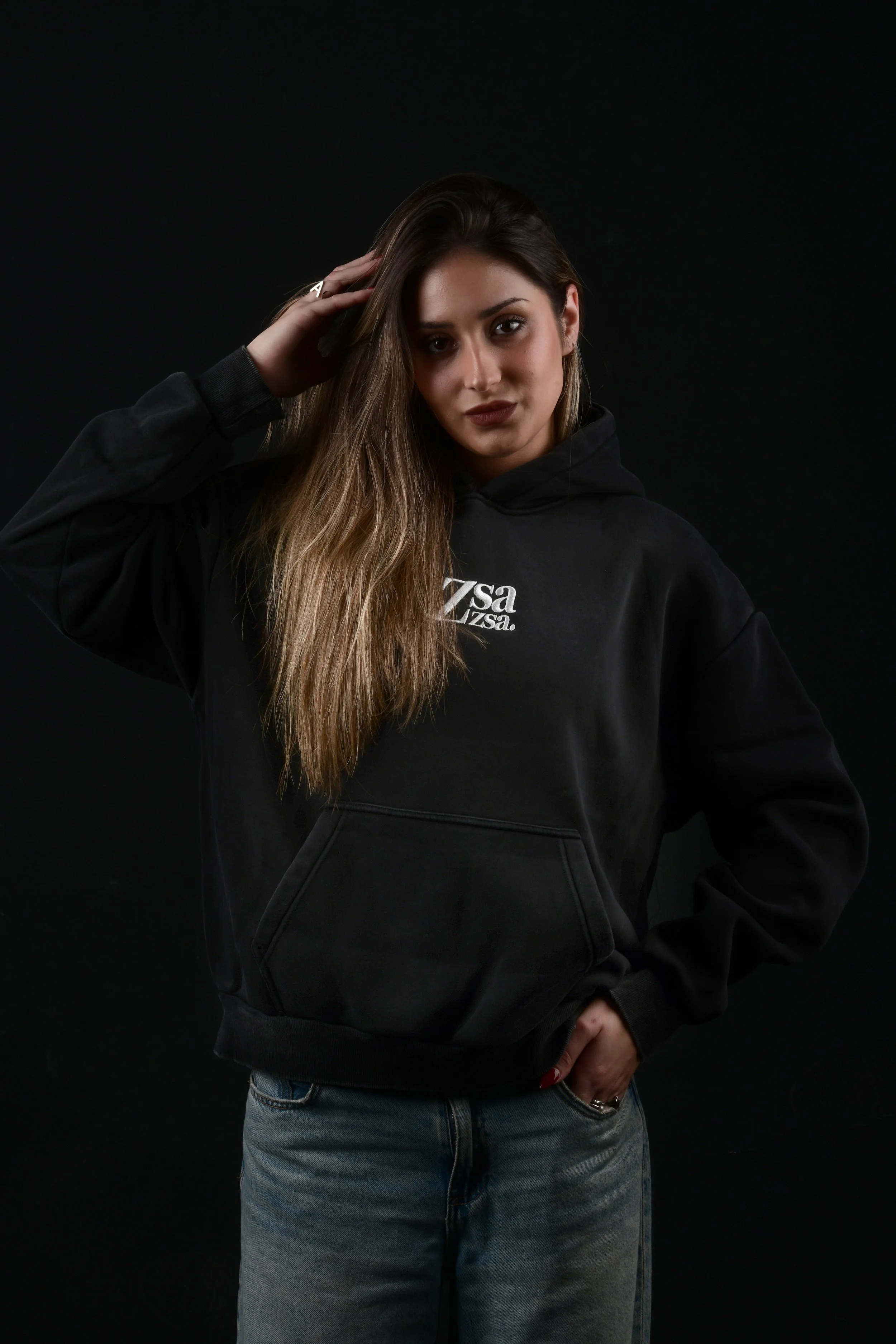 Juventud mujer con cabello largo y castaño claro, vestida con sudadera negra y jeans, posa contra fondo negro.
