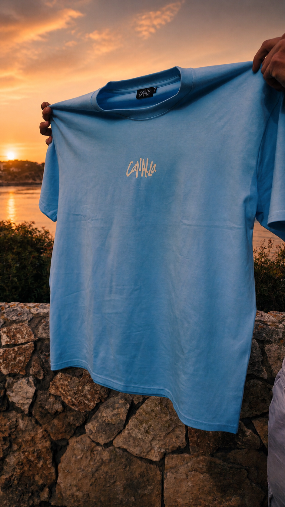 Playera azul con la palabra "Cayulla" en el centro, sostenida por una persona frente a un atardecer sobre un río y cerca de una pared de piedras.