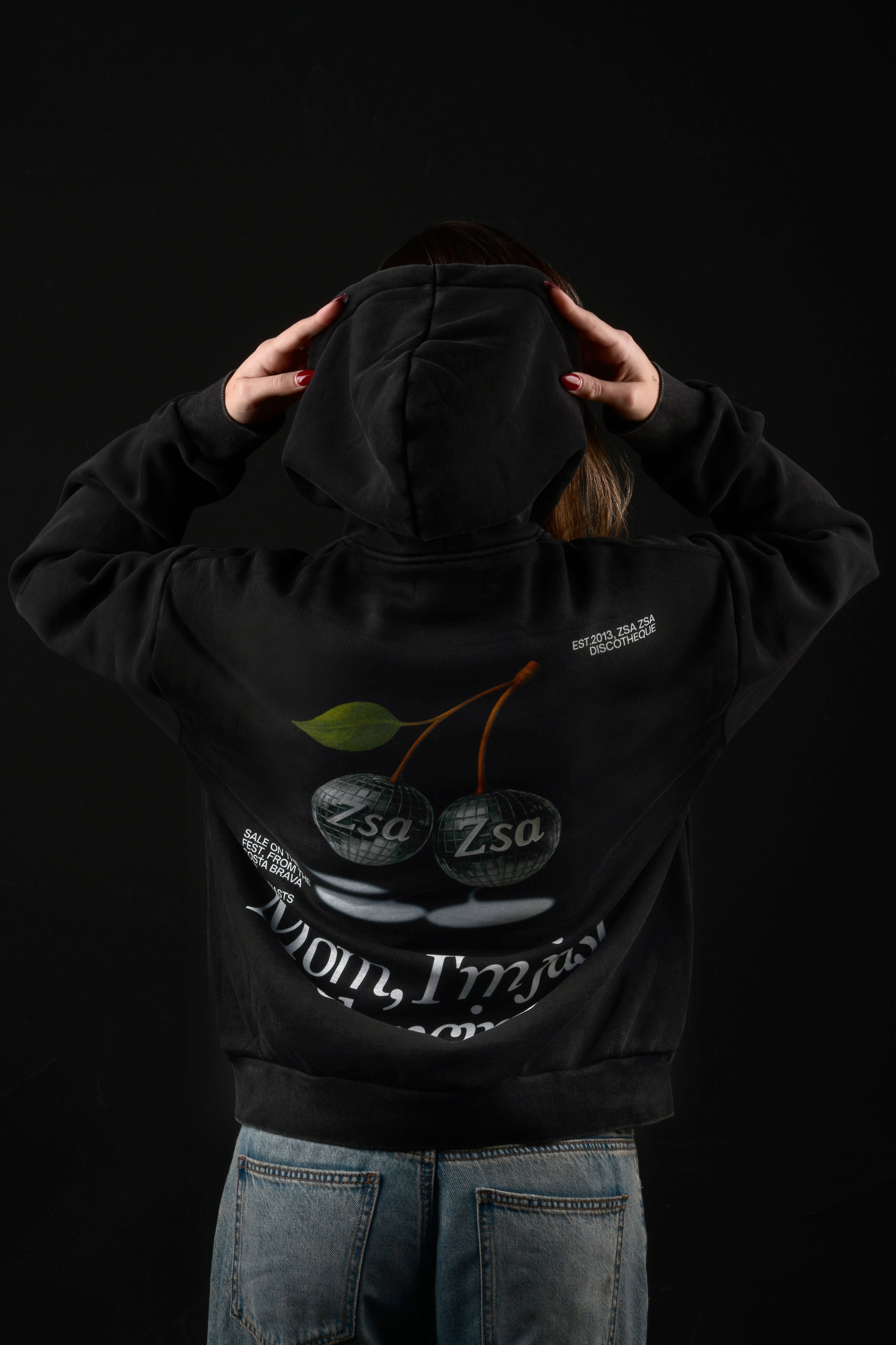 Persona vista desde atrás con sudadera negra con capucha, jeans claros, y sosteniendo una capucha negra. La sudadera tiene un diseño con varios globos con el texto 'Zsa' y letras adicionales, en un fondo negro.