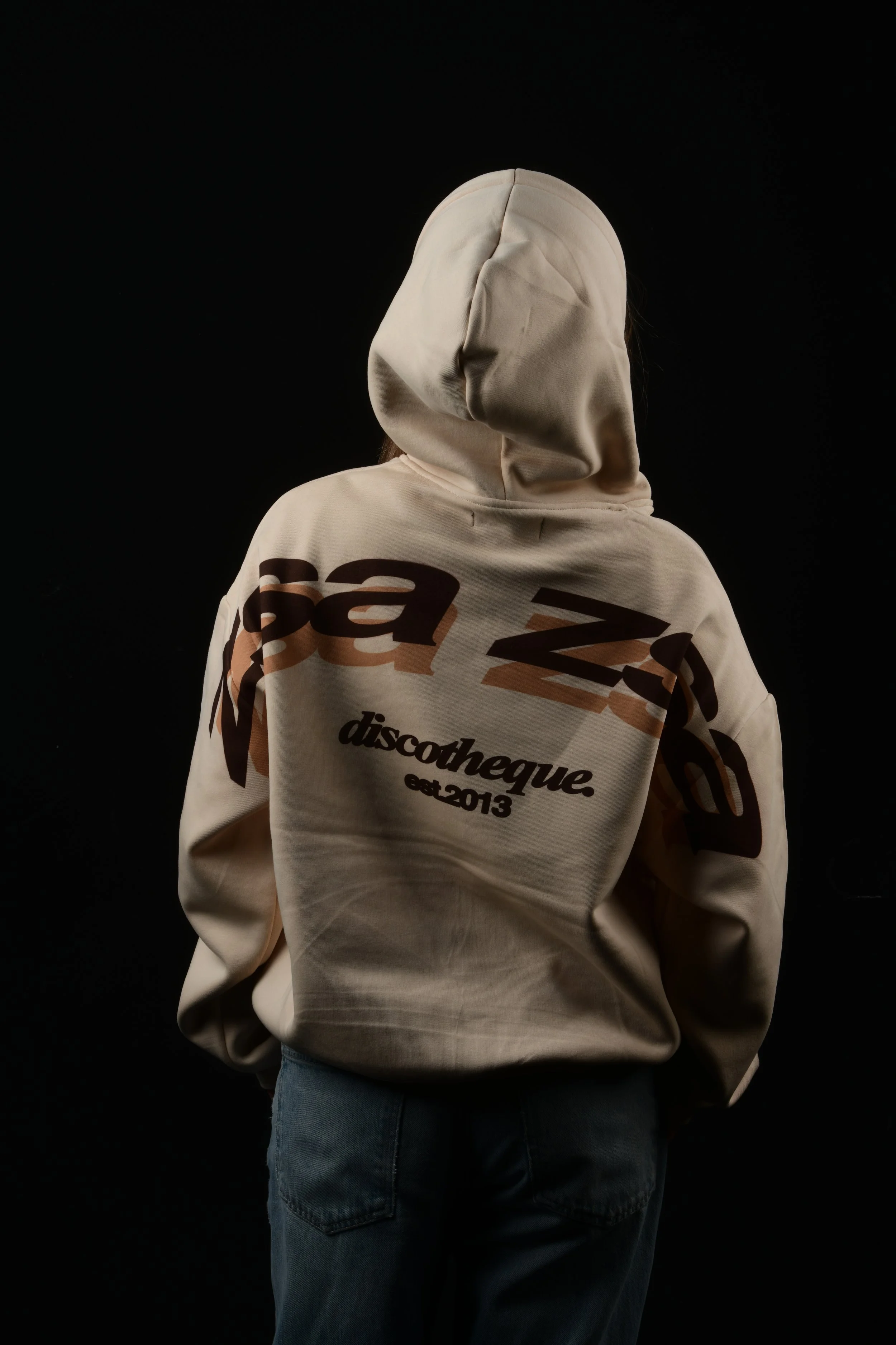 Persona con sudadera beige con letras grandes y oscuras, pantalón jeans azul, de espaldas, contra fondo negro.