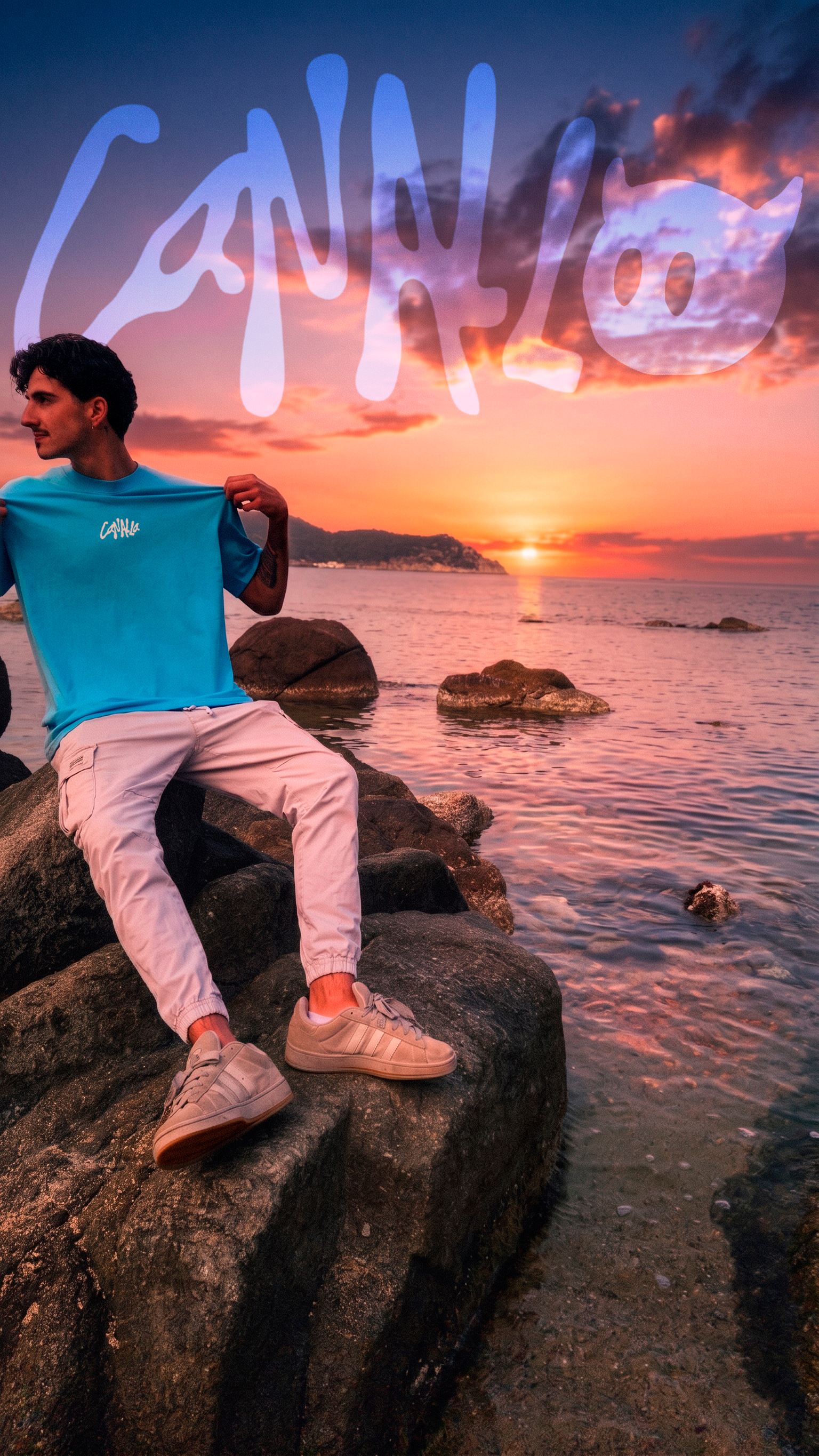 Un hombre con camiseta azul y pantalones blancos sentado en rocas junto al mar al atardecer