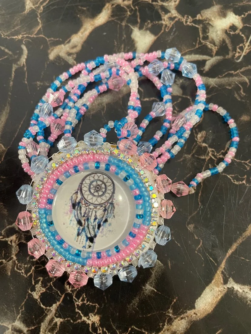 A beaded necklace with a dreamcatcher pendant featuring a cat illustration, decorated with pink, blue, and clear beads, resting on a black marble surface.