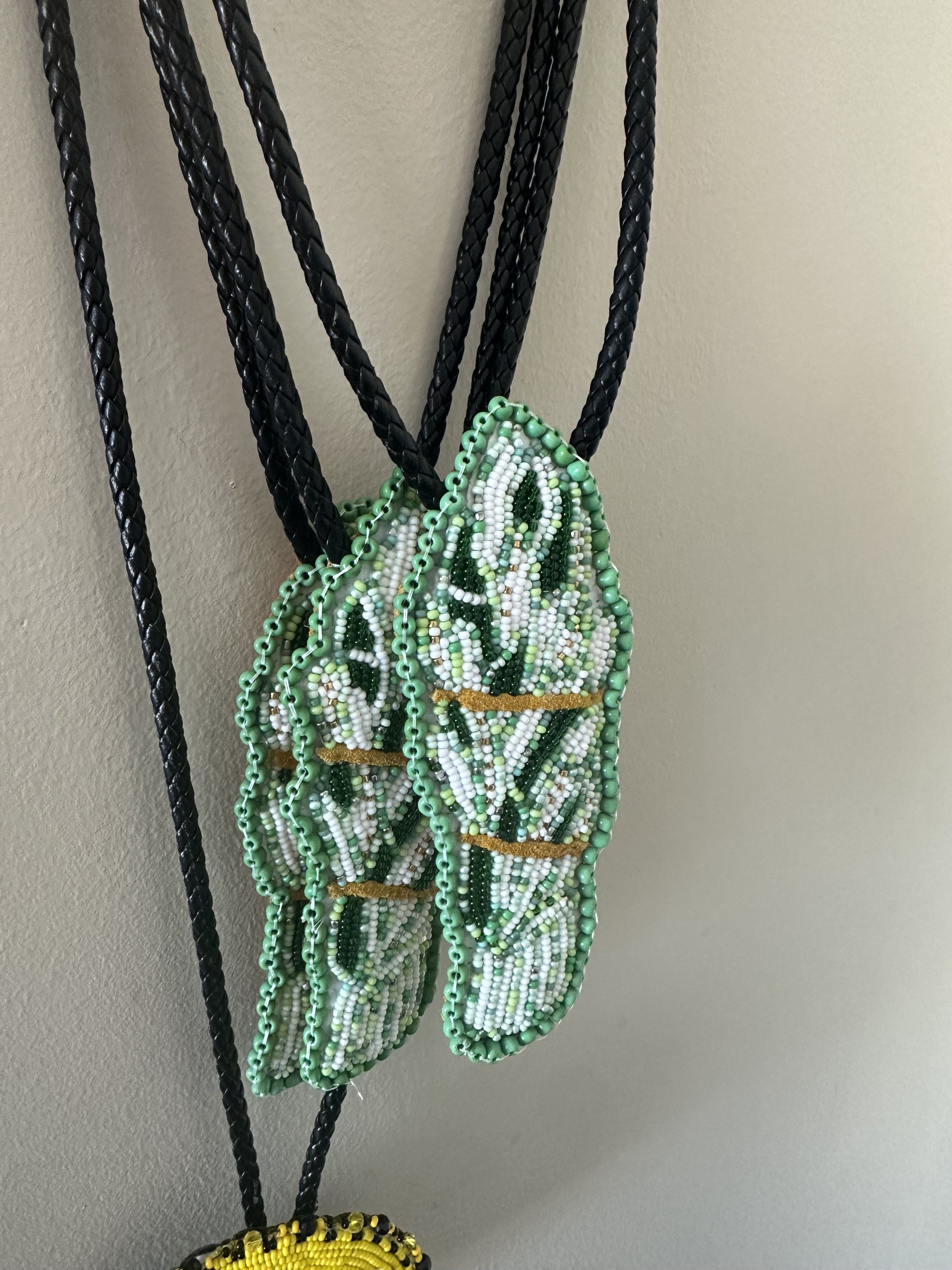 Beaded jewelry necklaces hanging against a beige wall. The necklaces include a turquoise, white, and green beaded piece designed like a butterfly, and a yellow and black circular beaded piece.