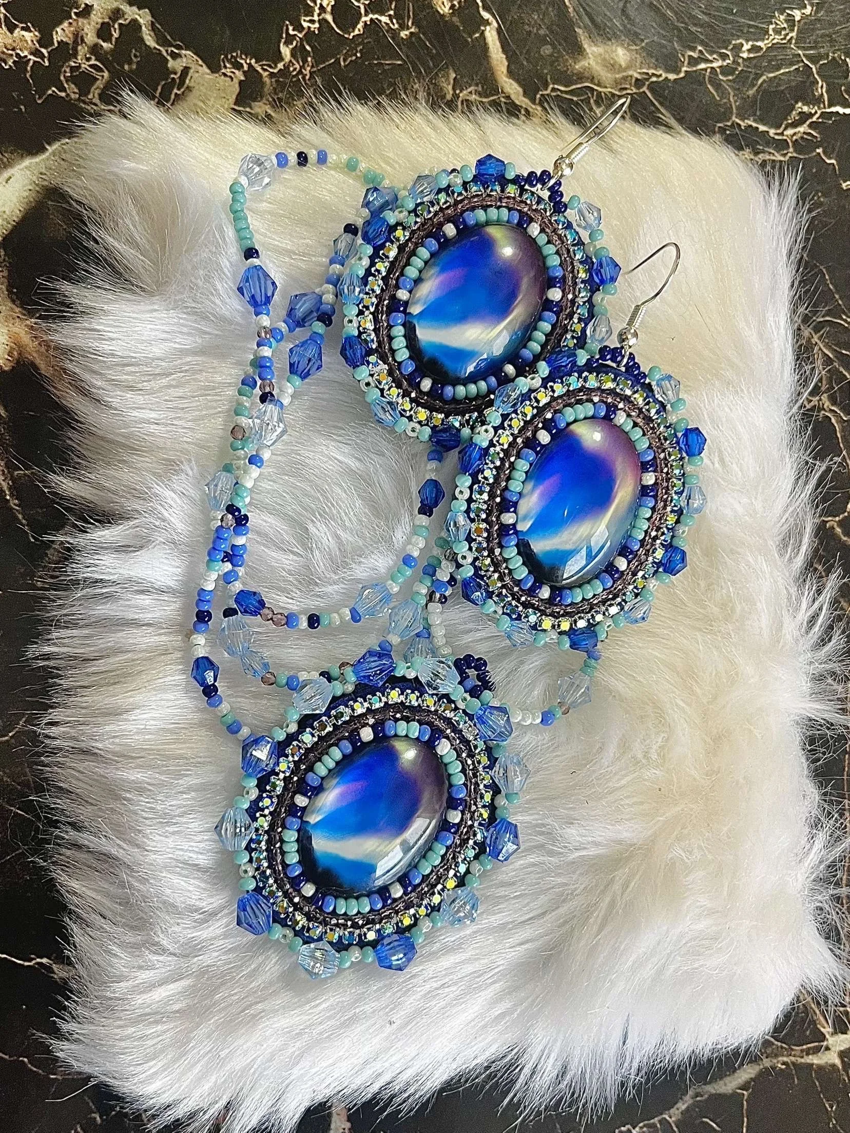 A pair of beaded earrings with large oval blue and purple cabochon stones, surrounded by small blue, turquoise, white, and clear beads, set on white faux fur with a dark marble surface background.