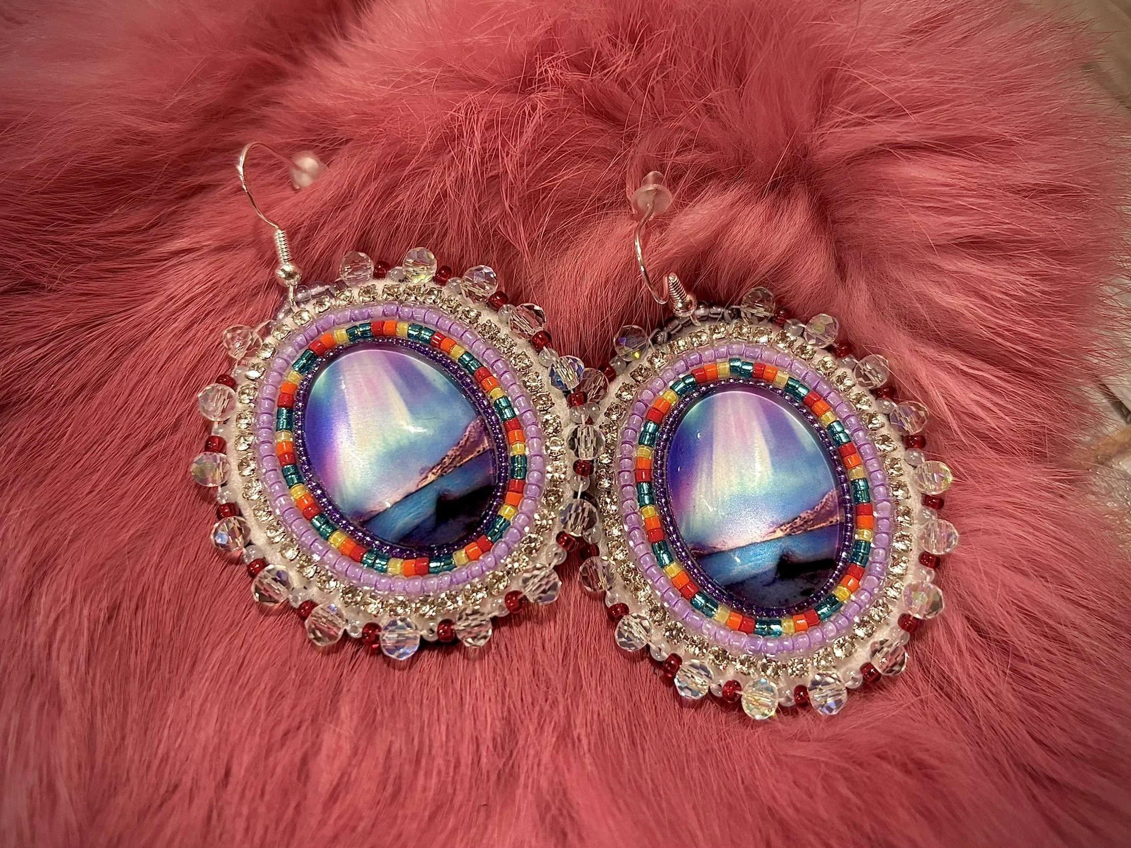 Colorful beaded earrings with mirrored centers, surrounded by rhinestones and beads, on pink furry fabric.
