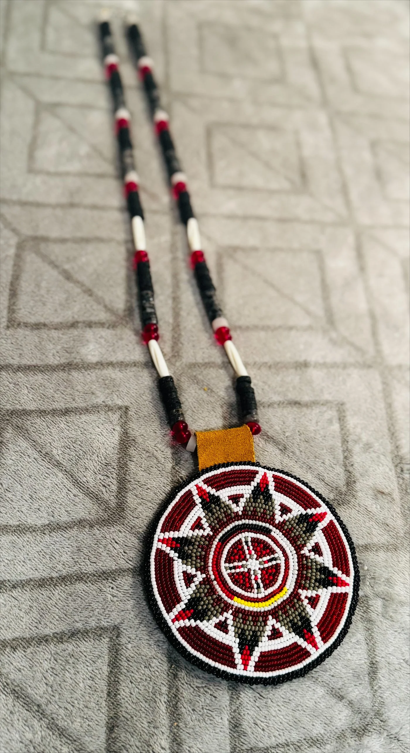 A beaded necklace with black, white, red, and gray beads, featuring a circular pendant with a red, black, white, and yellow pattern, resting on a textured gray surface.