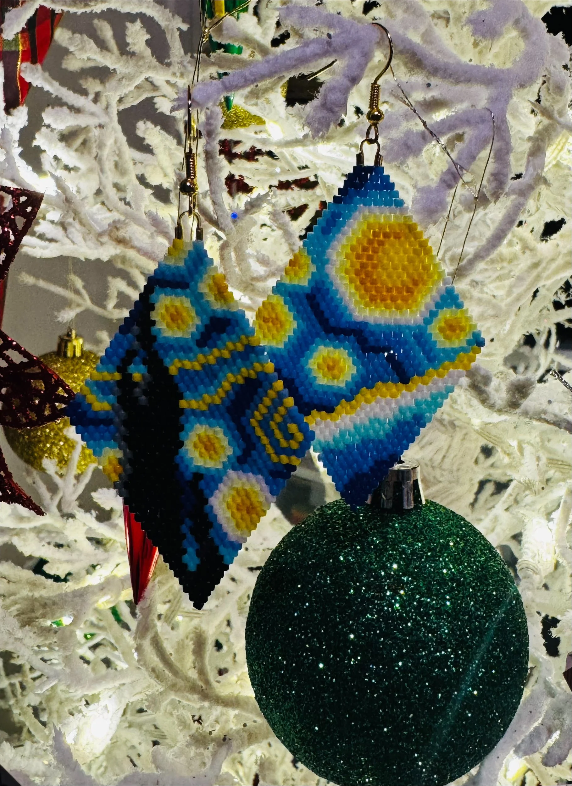 Ornaments hanging from a white decorated Christmas tree, including a beaded butterfly with a blue and yellow pattern, a large green glittery ball, and a smaller gold ornament.