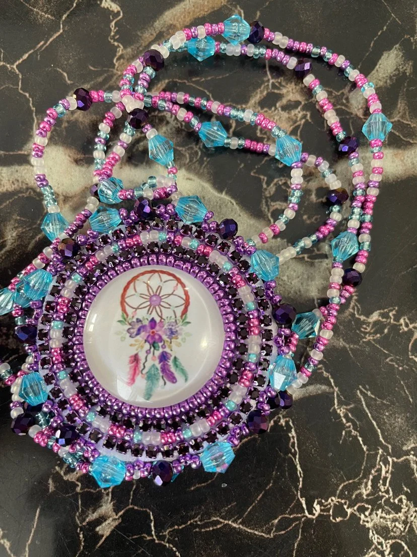 Colorful beaded necklace with purple, pink, white, and blue beads, centered around a circular pendant with a dreamcatcher and floral design, laid on a black marble surface.