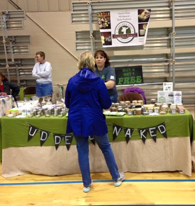 A vendor booth at a market selling handmade natural body products. A banner reads "Udderly Naked," and jars are displayed on the table. A person is interacting with the vendor.