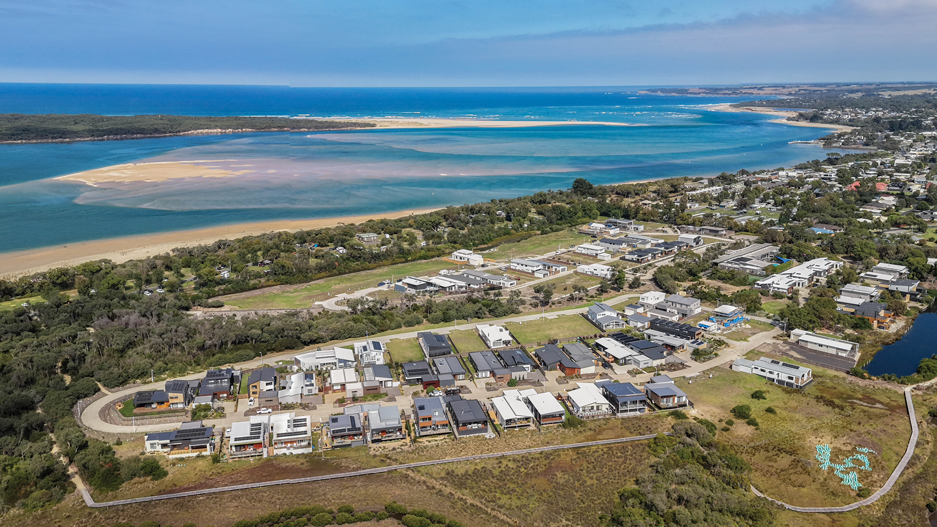 Aerial view of a coastal area with a beach, water body, and nearby greenery. There are buildings and a road near the shore. The image shows a mix of natural landscape and developed areas under a clear blue sky.