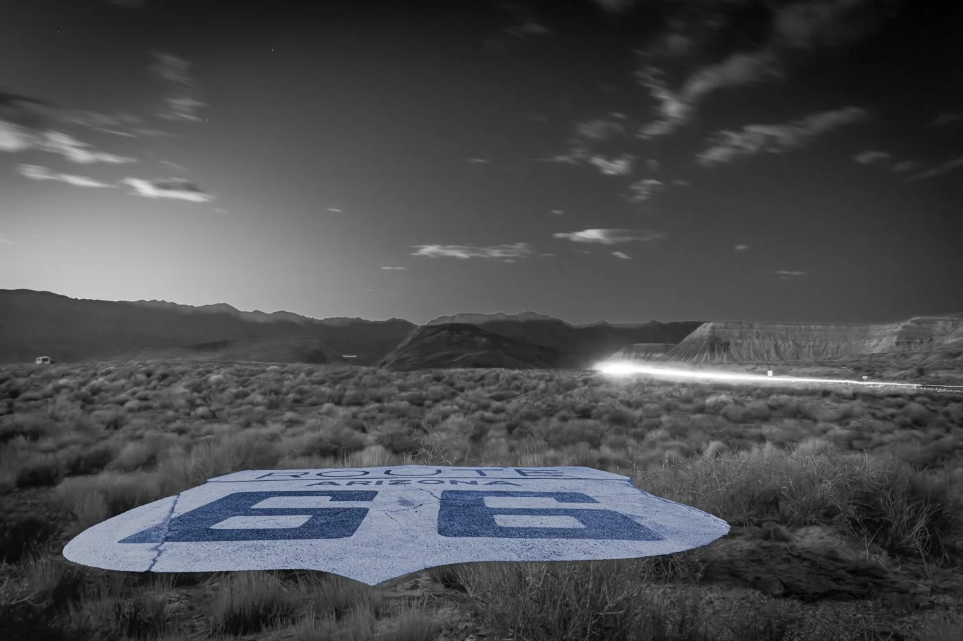 Black and white fine art photograph of a Route 66 shield painted on the road with a car light trail passing by.