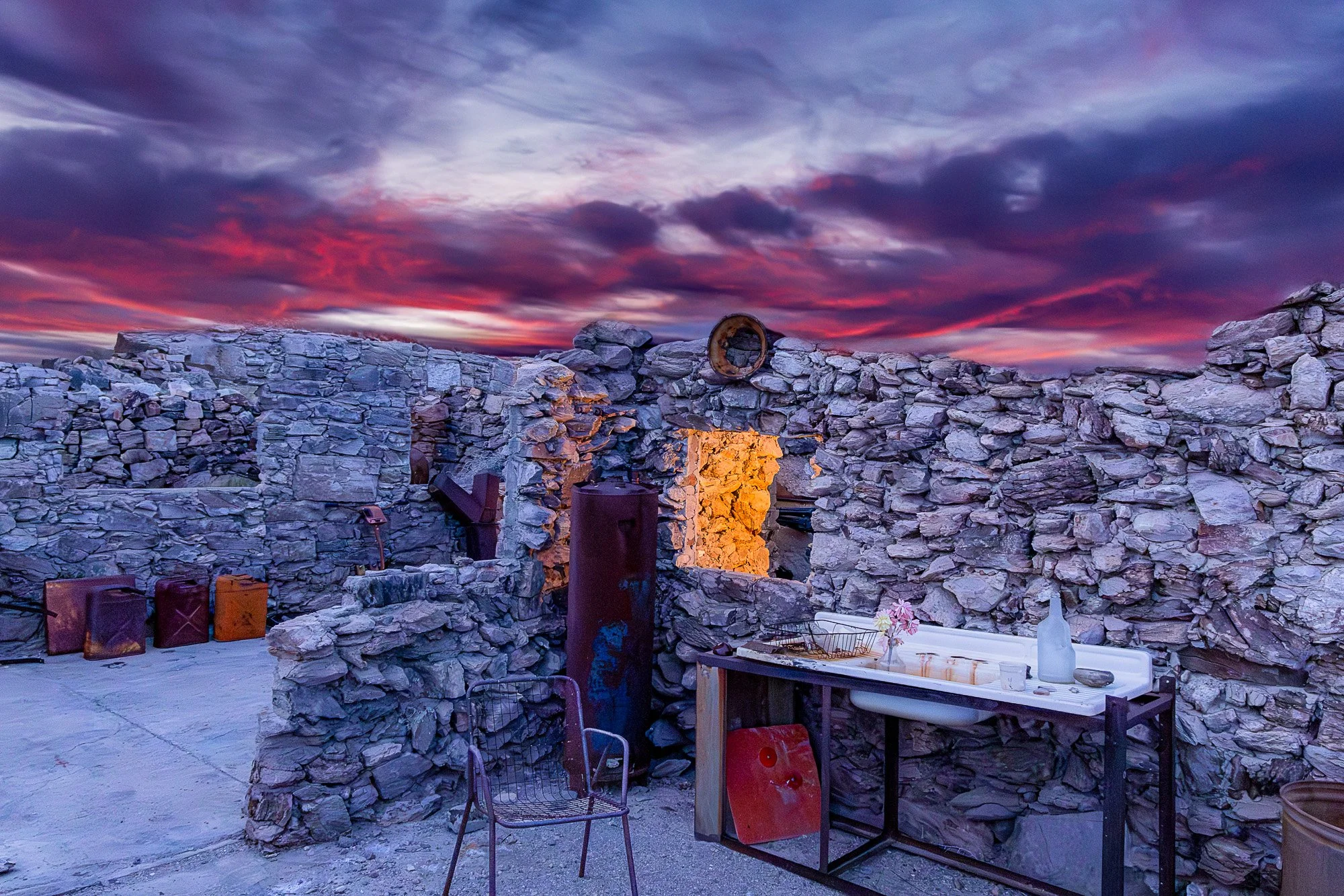 Outdoor scene at sunset with a stone wall, tables, chairs, and various objects under a colorful sky. Gold Eye Mine, AZ