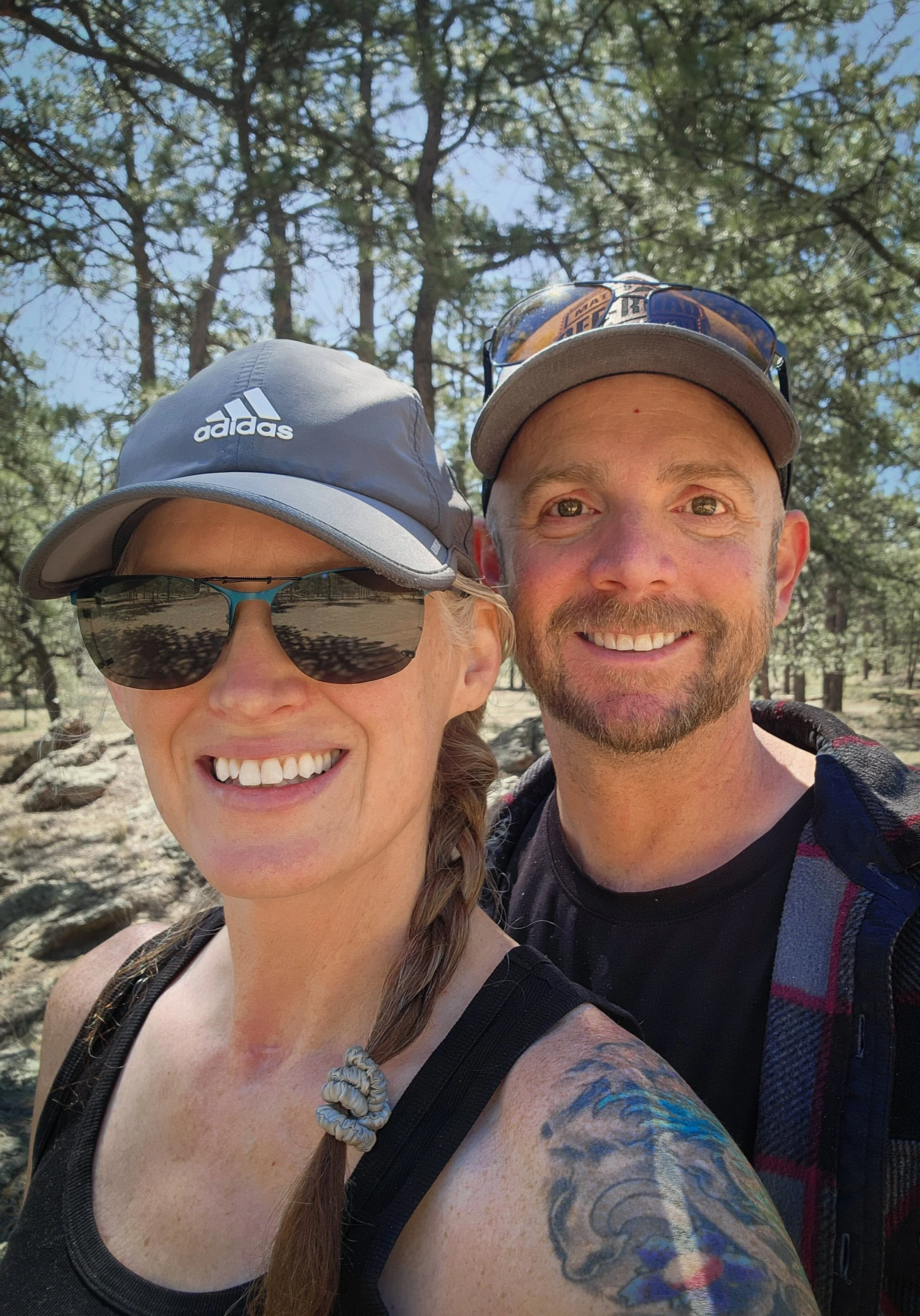 Two people outdoors in a forested area taking a selfie. One woman with sunglasses, a gray Adidas cap, and a braid, smiling. One man with a cap and sunglasses on his cap, smiling. Tall pine trees in the background.