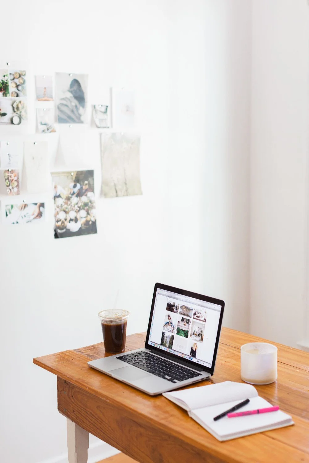 A wooden desk with a laptop displaying a photo website, a glass of iced coffee, an open notebook with pink and black pens, and a lamp. A white wall behind the desk features a collage of photographs.