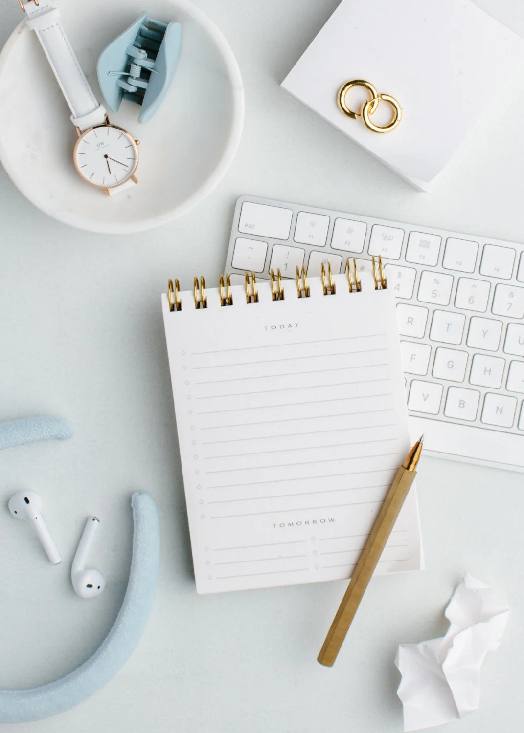 Flat lay of a white desk with a white keyboard, a spiral notepad with 'Today' and 'Tomorrow' sections, a gold pen, a white watch, gold earrings, light blue hair clips, white earbuds, and a crumpled piece of paper.