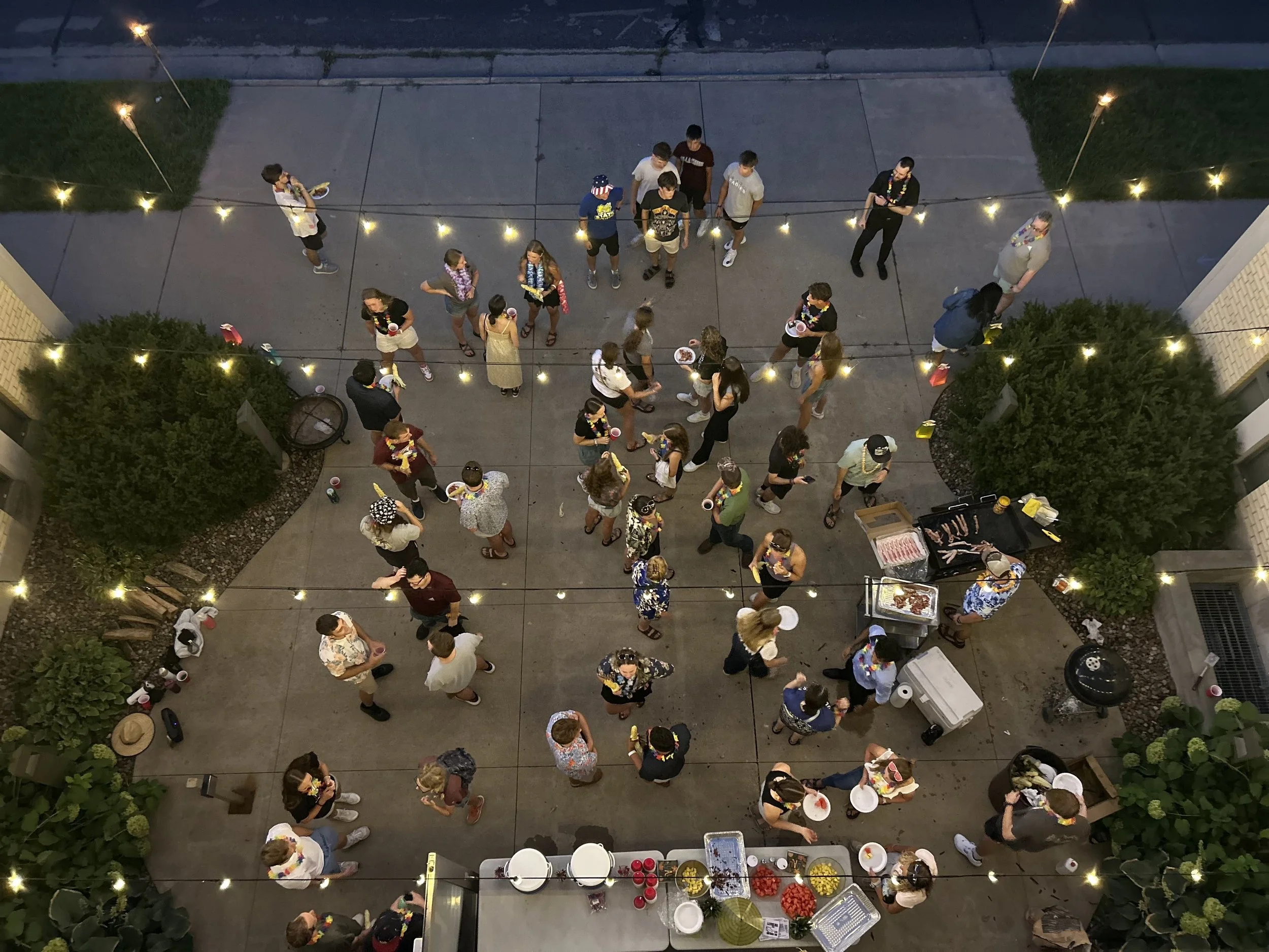 Overhead view of a group of people at an outdoor party, some wearing leis and casual summer clothing, gathered in a courtyard decorated with string lights, with a table of food and drinks nearby.