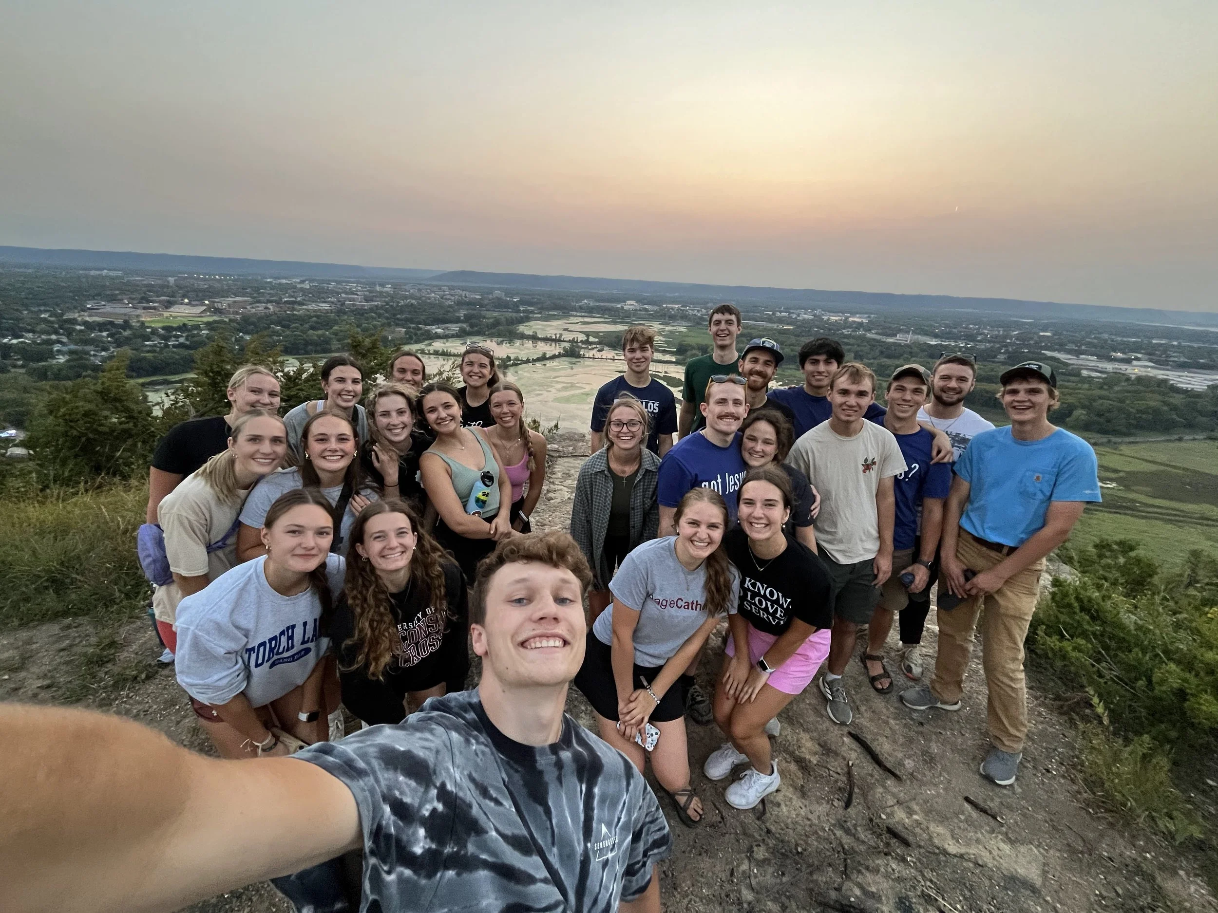 Group of smiling young people taking a selfie on a hilltop during sunset, overlooking a scenic landscape of river, greenery, and distant city.