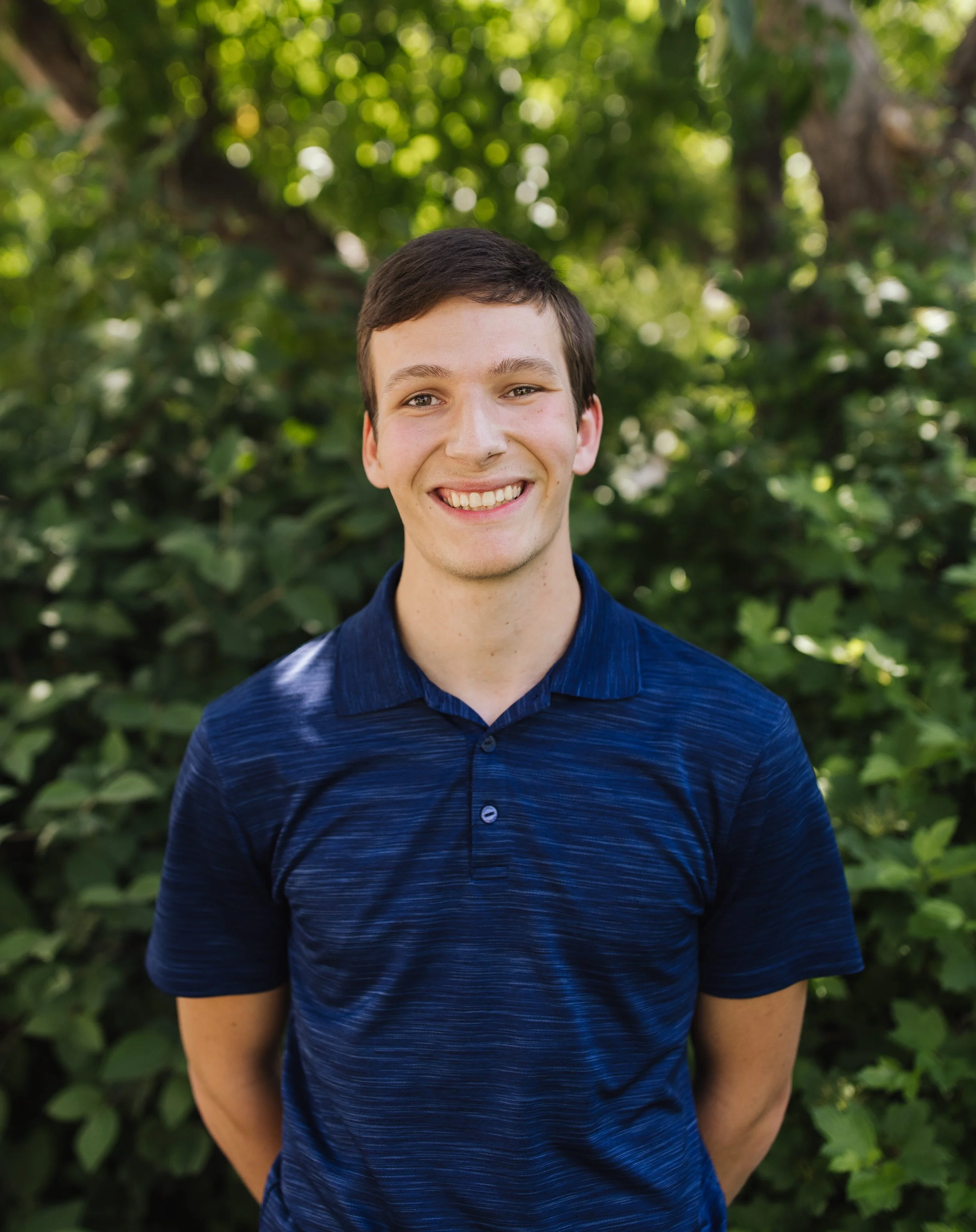 A young man with short dark hair and a big smile, wearing a blue polo shirt, standing outdoors in front of a green leafy background.