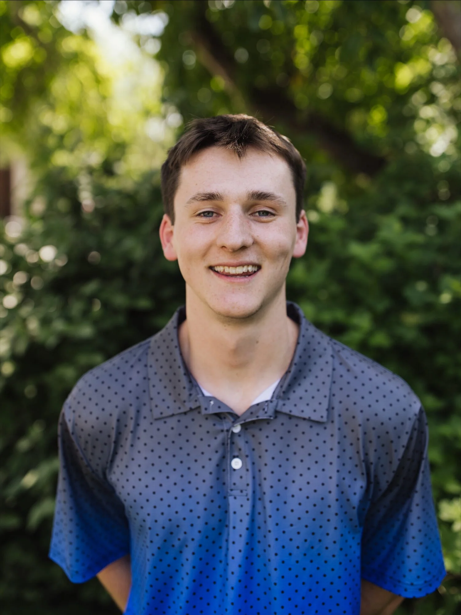 A young man with short brown hair and blue eyes smiling outdoors, with green trees and foliage in the background.