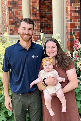 John & Caitlin Collins smiling while holding a baby girl in front of a brick building and colorful flowers.