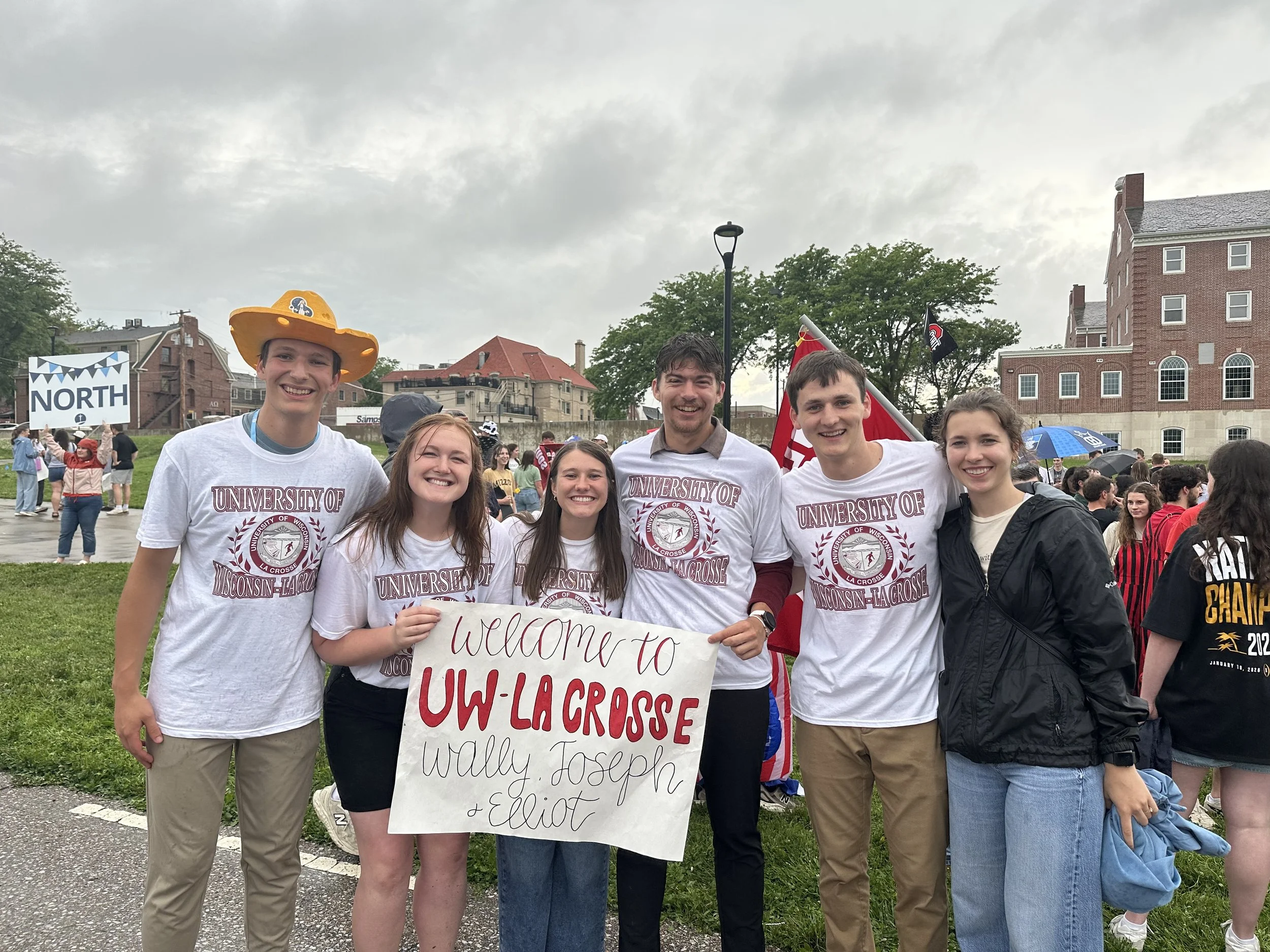 A group of people at an outdoor event, holding a sign about UW-Lacrosse lacrosse, with some dressed in university apparel, and others wearing casual clothes. The background shows more people, flags, and historic buildings under a cloudy sky.