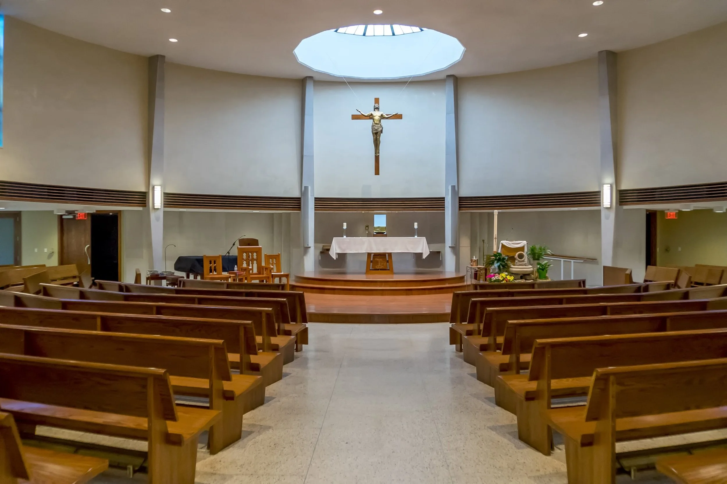 Interior of a church sanctuary with wooden pews, an altar, a crucifix hanging on the wall above the altar, and a skylight overhead.