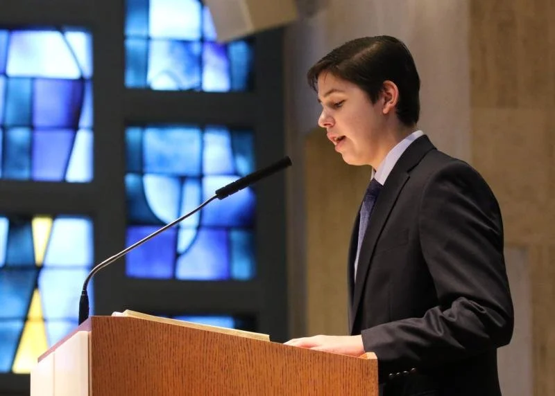 A young man in a dark suit and tie speaking at a podium in front of a colorful stained glass window.