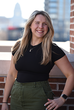 Eliese with long blonde hair wearing a black t-shirt and green pants, standing outdoors near a brick wall with a cityscape background.