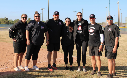 Group of people standing on a baseball field wearing matching team shirts and caps, posing for a photo.