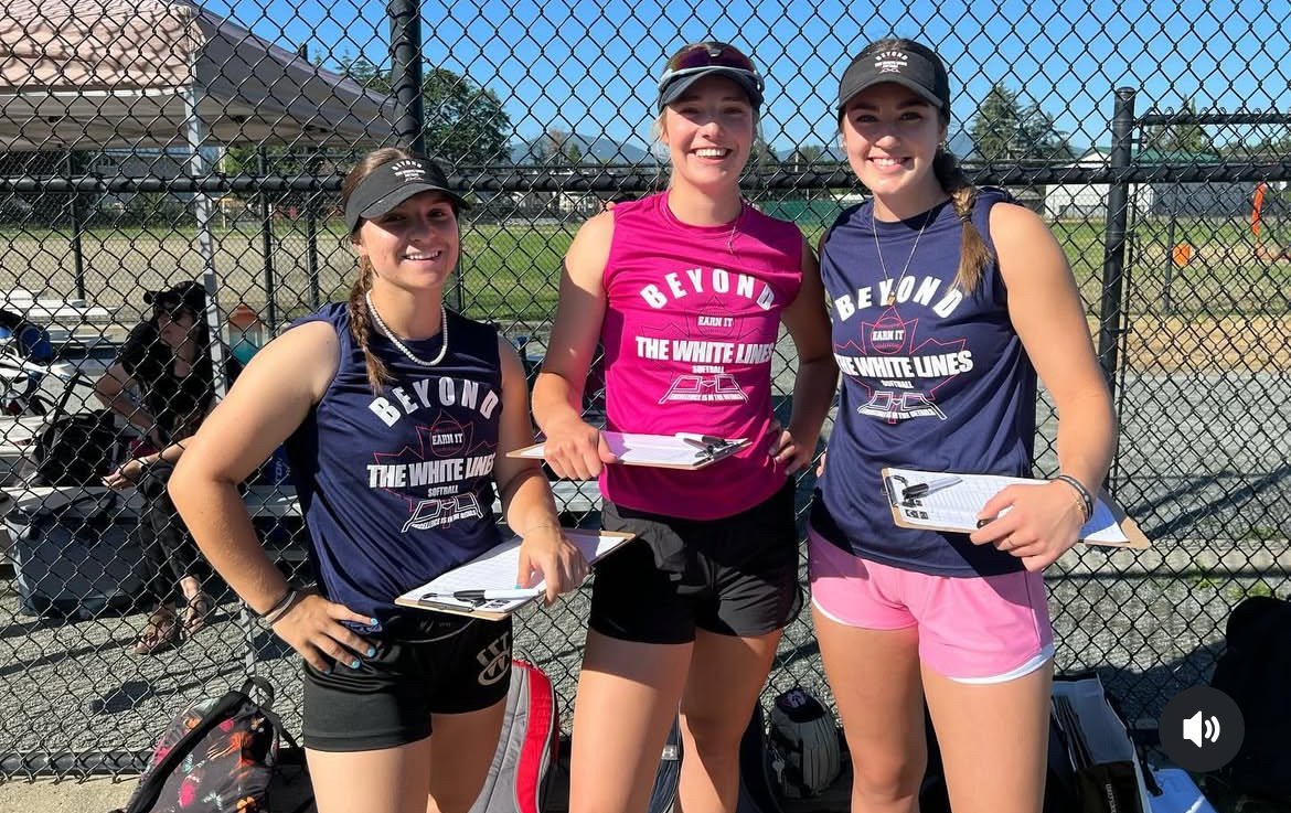Three women in athletic clothing with "Beyond the White Lines" text on their shirts, holding clipboards, standing in front of a chain-link fence at an outdoor sports facility.