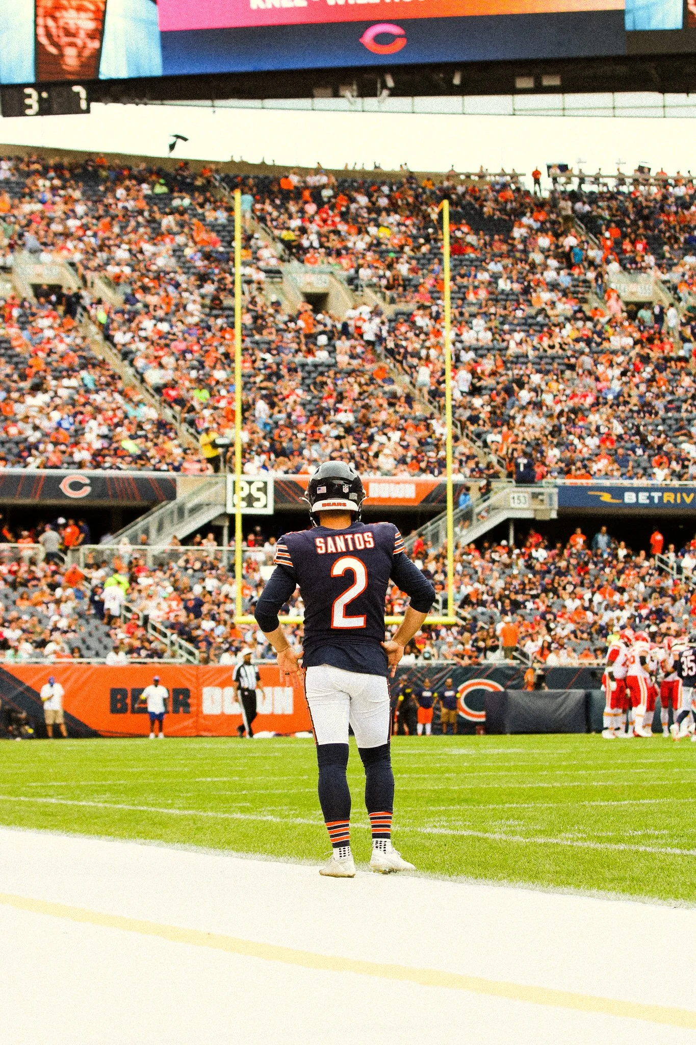 A football player wearing a Chicago Bears uniform with the name Santos and number 2 stands on the sideline during a game, with a crowded stadium in the background.
