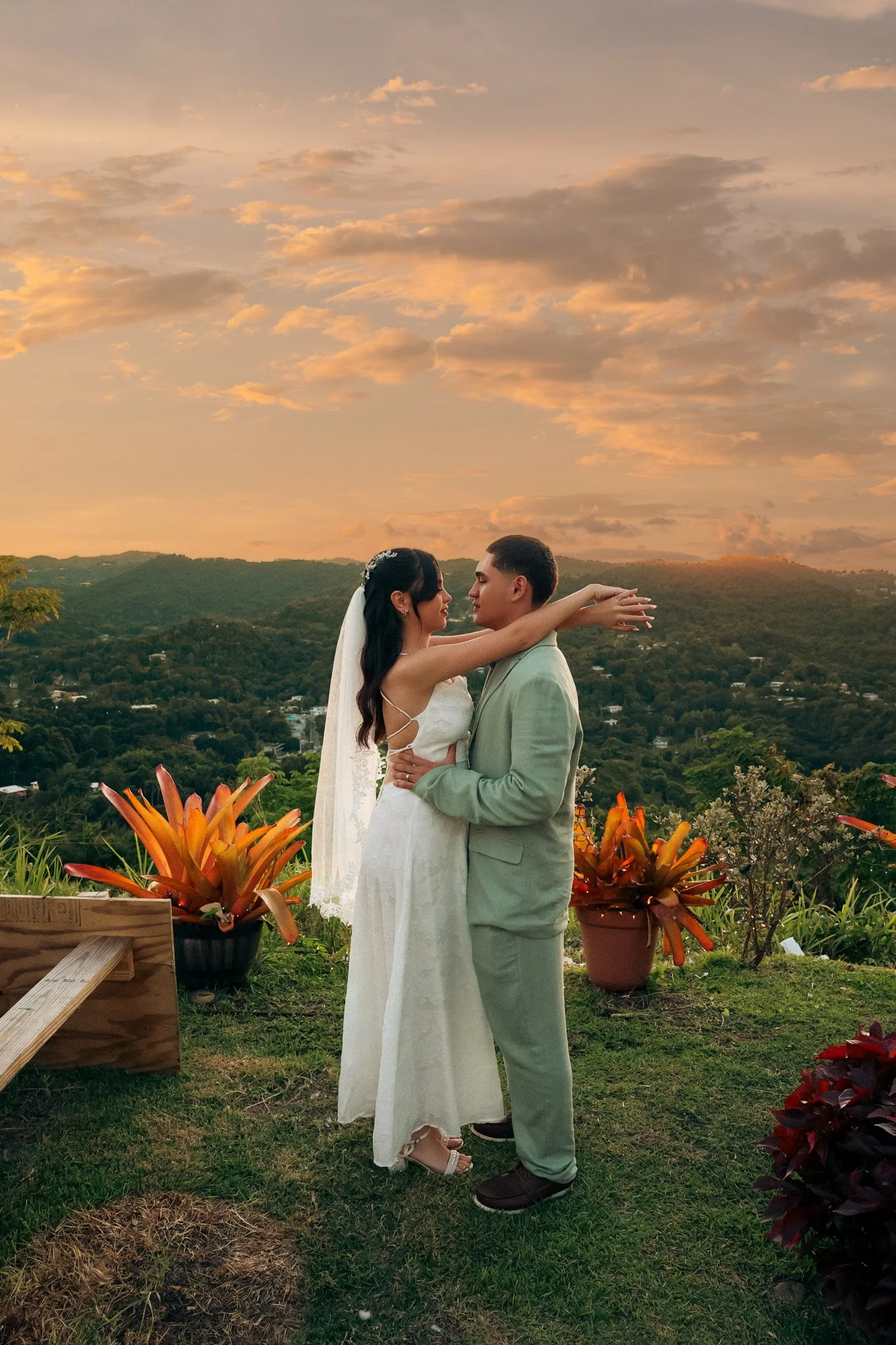 A bride and groom standing close together at sunset, embracing outdoors with hills in the background and potted plants around them.