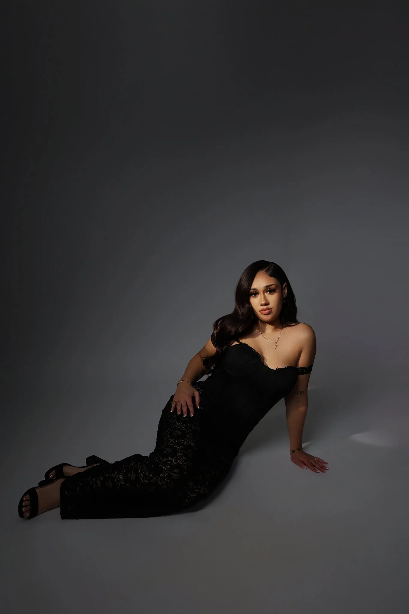 A woman in a black lace dress sitting on the floor against a dark gray background.