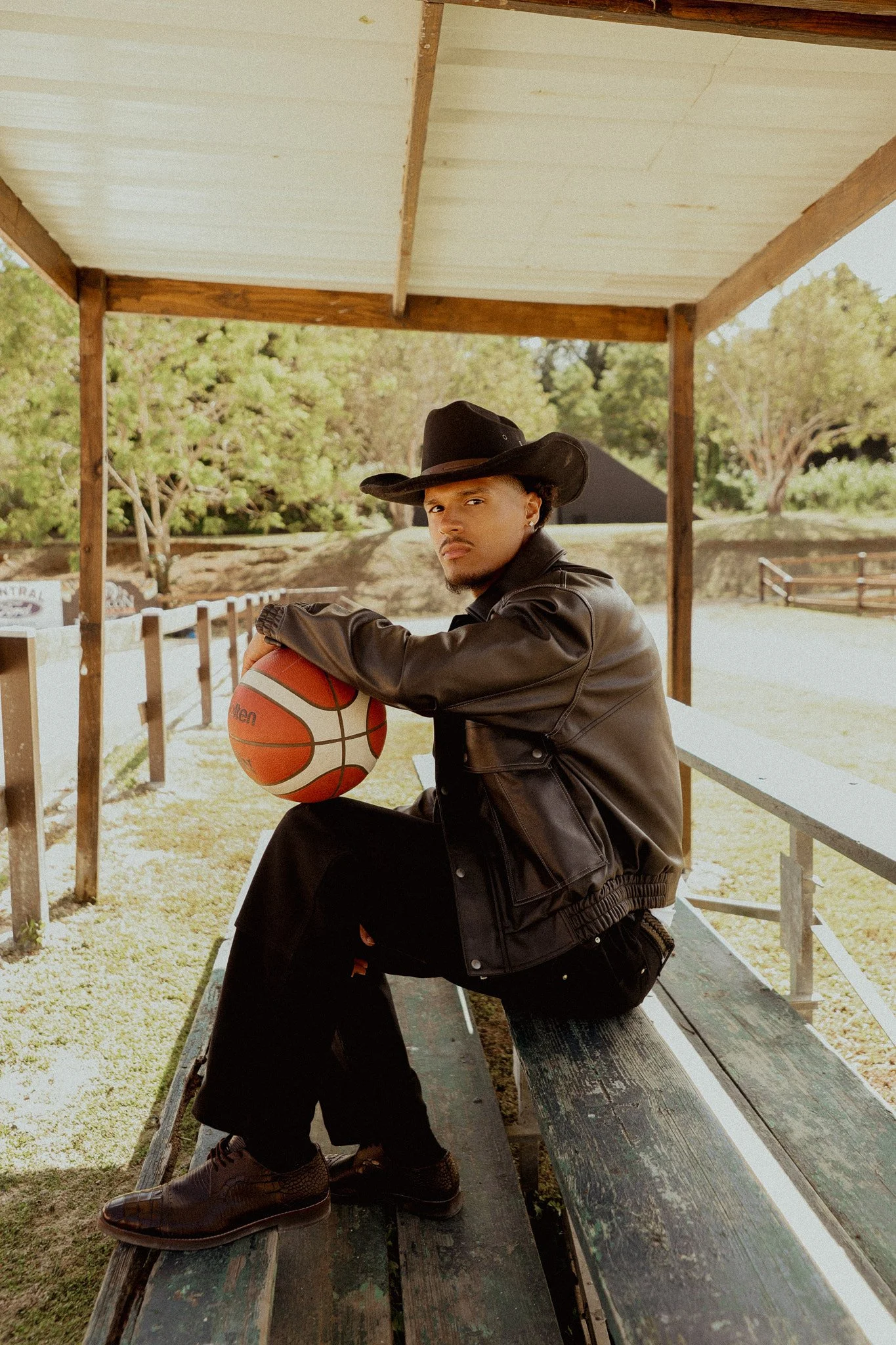 A young man wearing a black cowboy hat, leather jacket, and brown shoes sitting on a wooden bench, holding a basketball, outdoors with trees in the background, in Puerto Rico