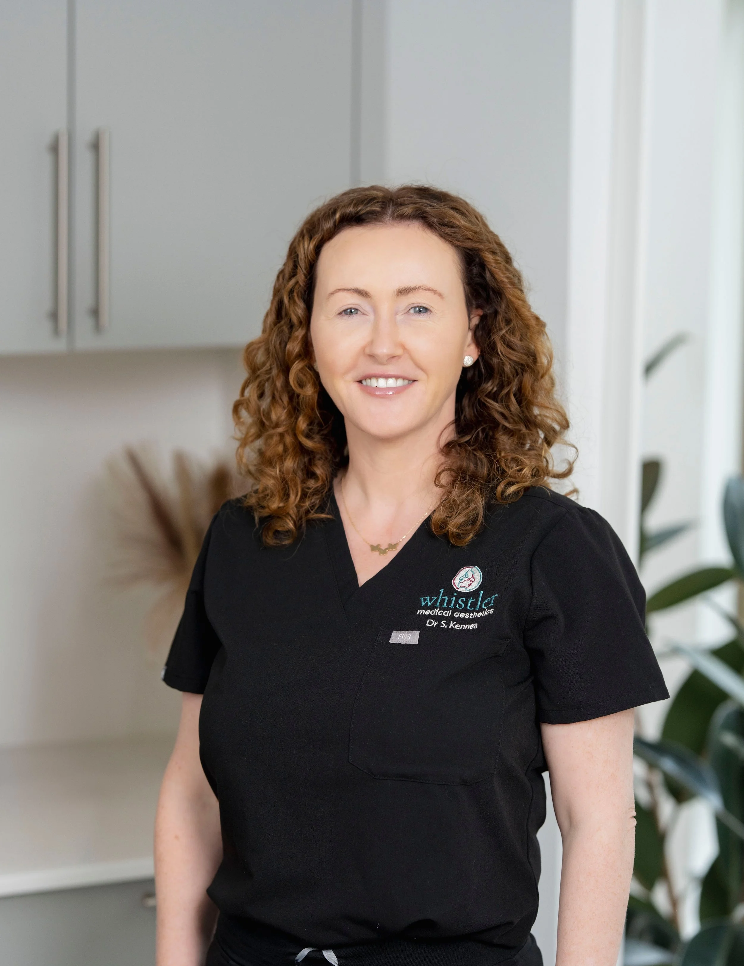 Person in black scrubs with curly hair standing indoors, smiling, with cabinets and plants in the background. Embroidered text on scrubs.