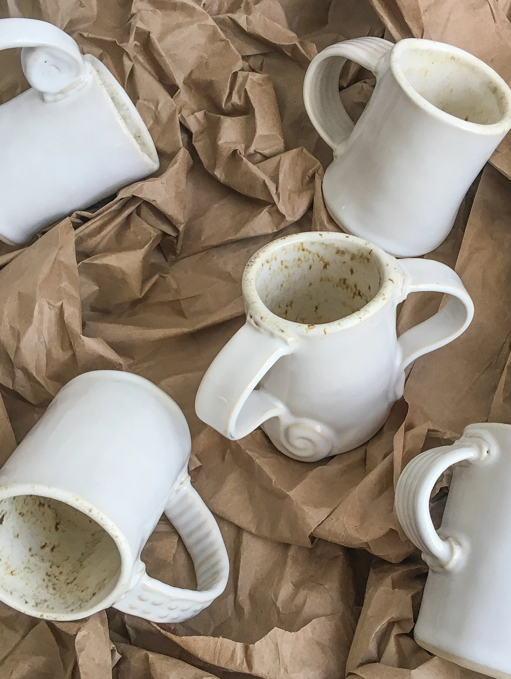 Multiple dirty white ceramic mugs with handles, some with lids, on crumpled brown paper.
